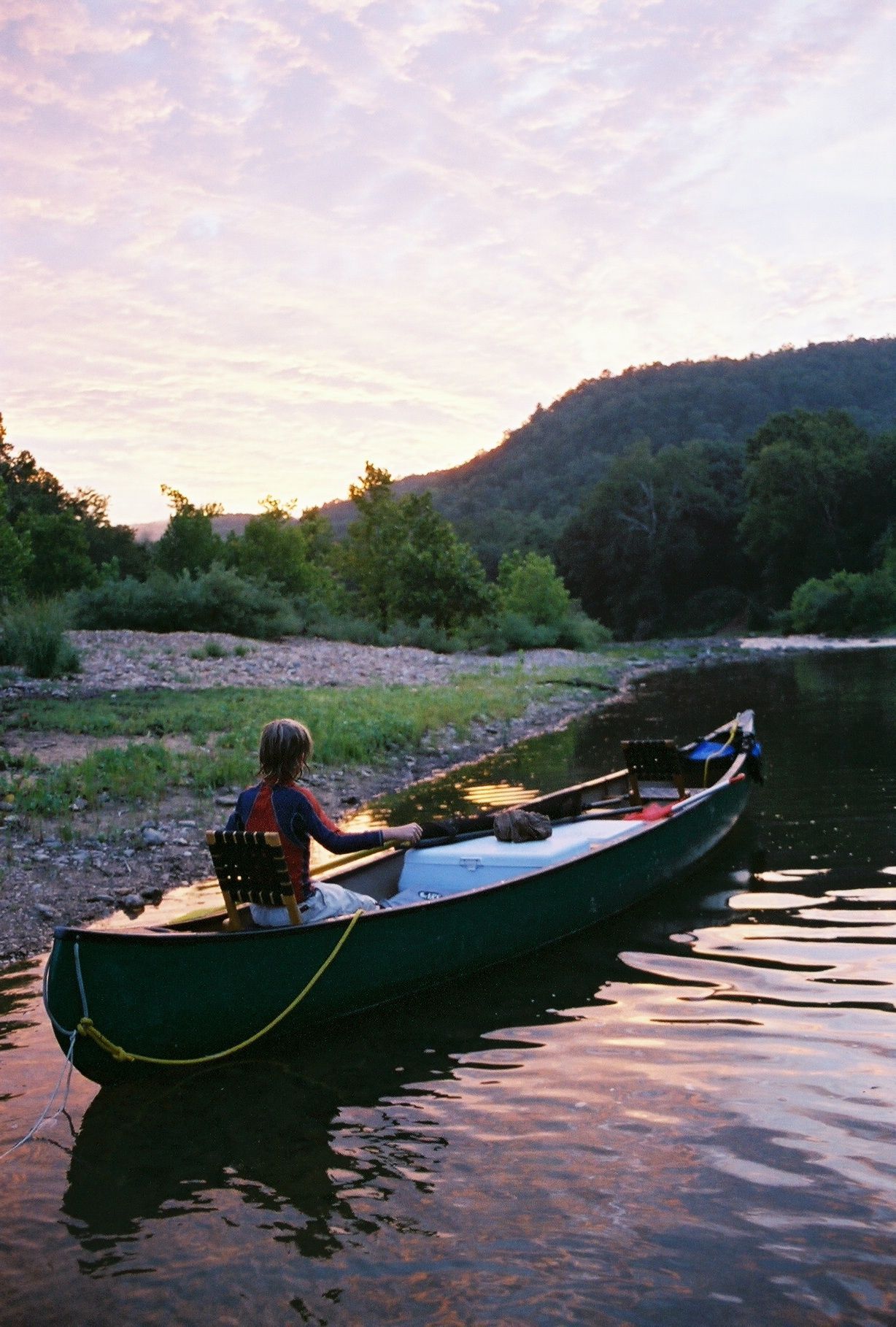 Person paddling a canoe on a calm river at sunset, with green hills and a pink sky.