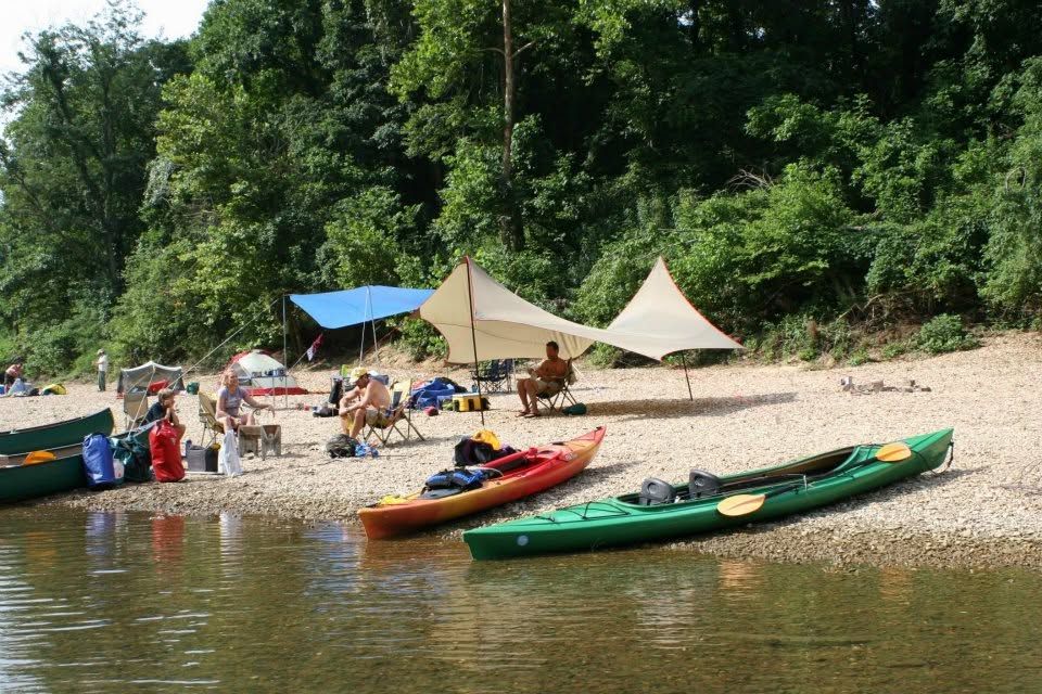 People lounging by colorful kayaks on a sandy riverbank under tents, with forested cliffs behind