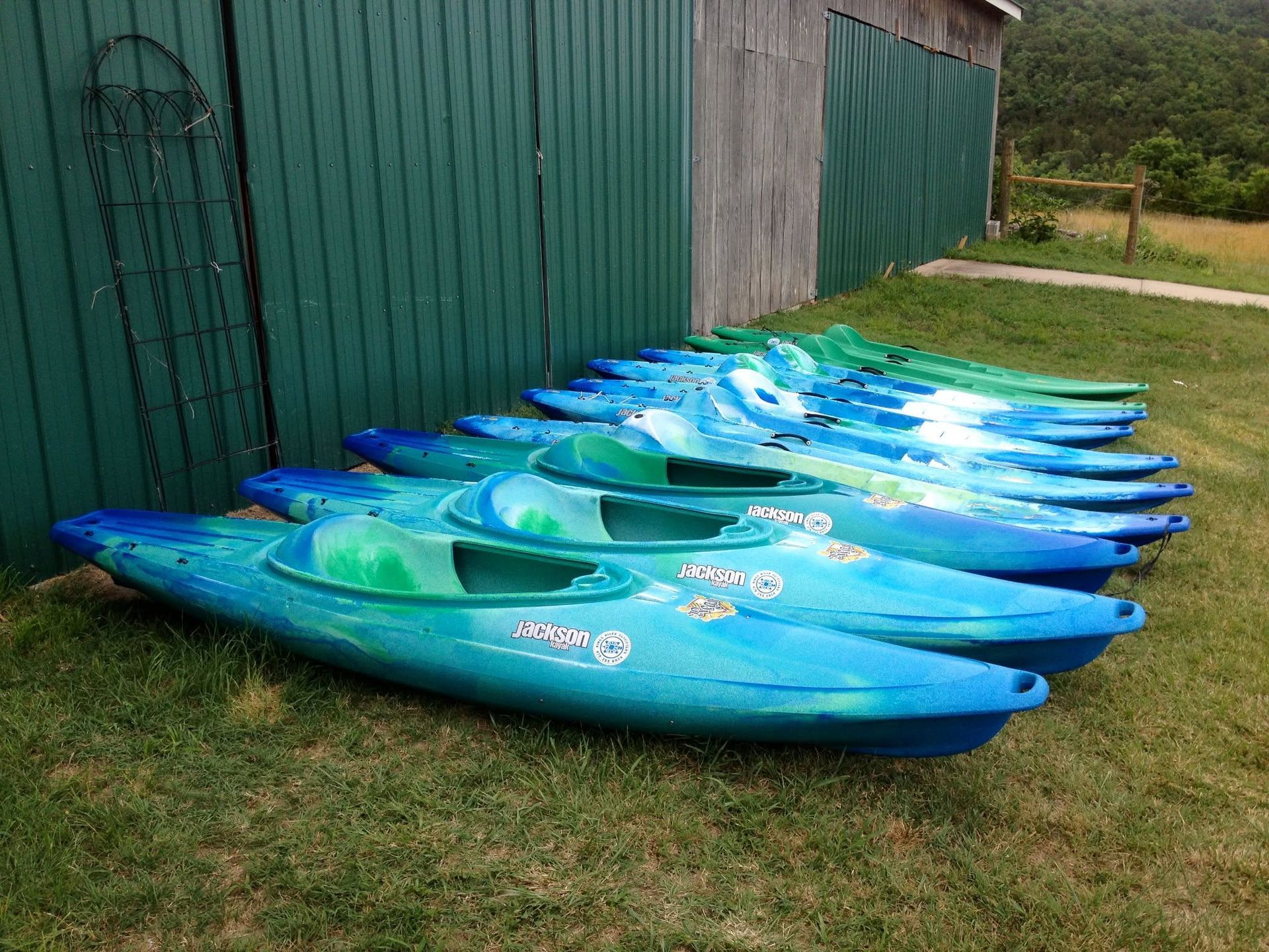 Row of blue kayaks stacked on grass beside a green shed outdoors