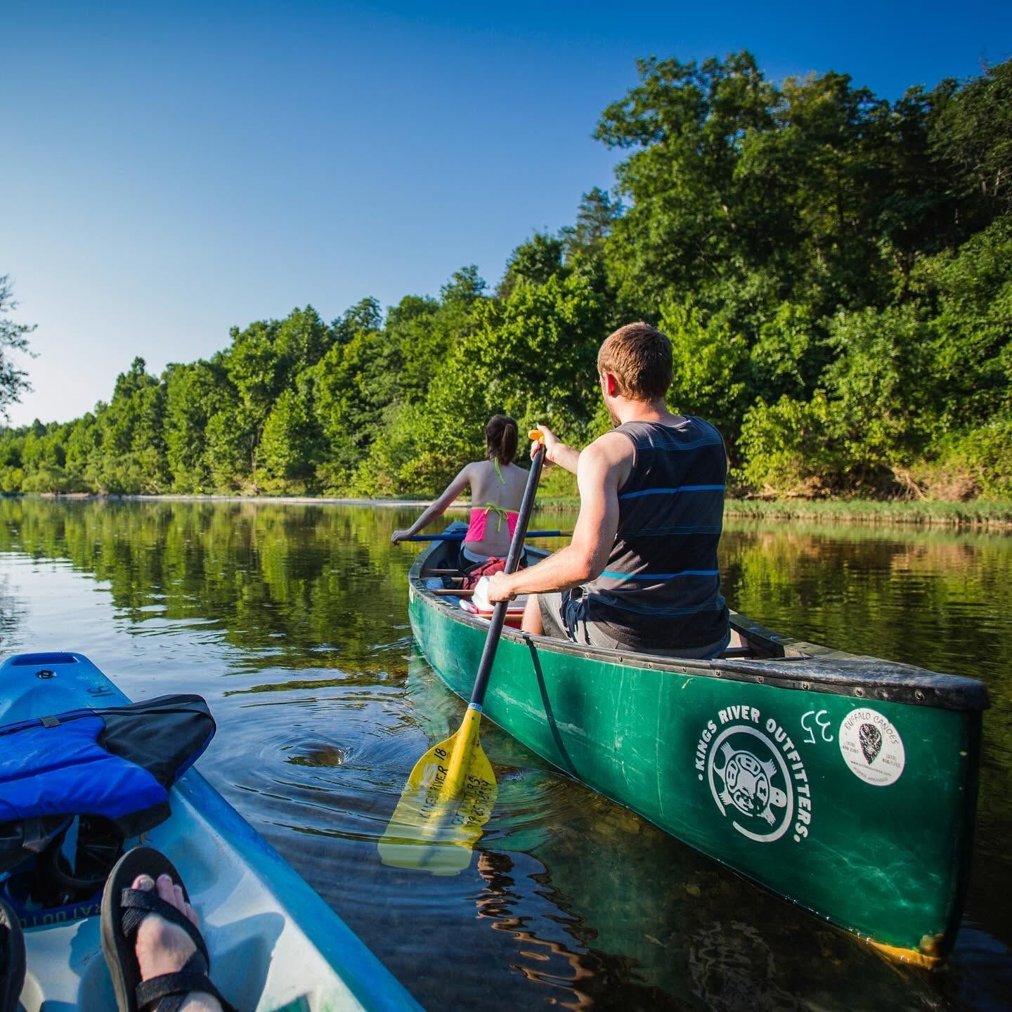 Two people kayaking on a calm river beside green trees under a blue sky
