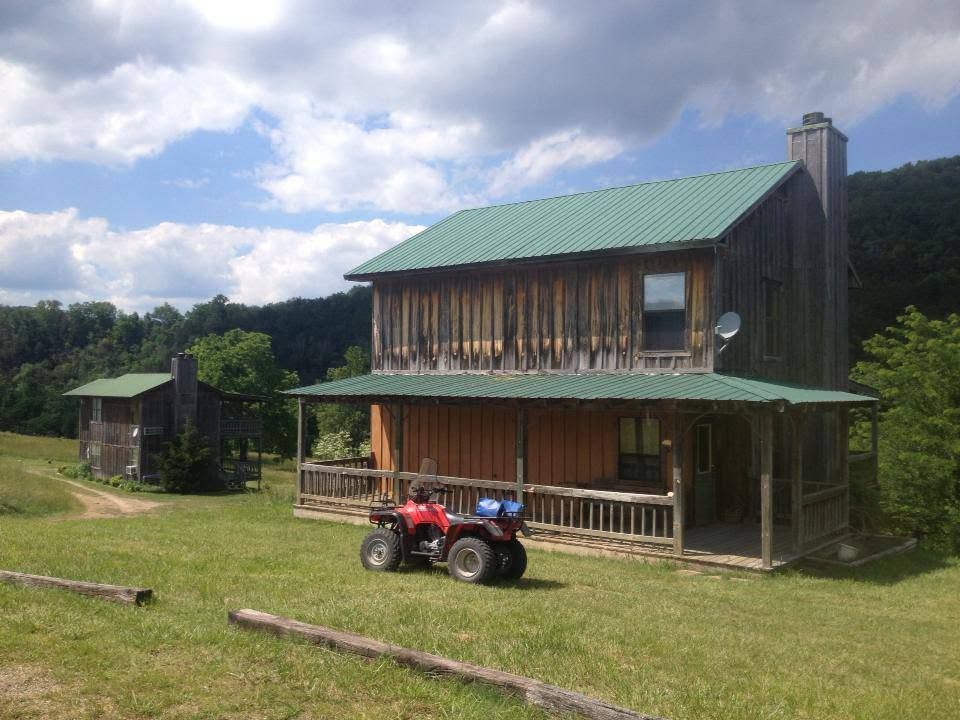 Rustic two-story cabin with green roof, porch, and red ATV in a grassy field under a cloudy sky
