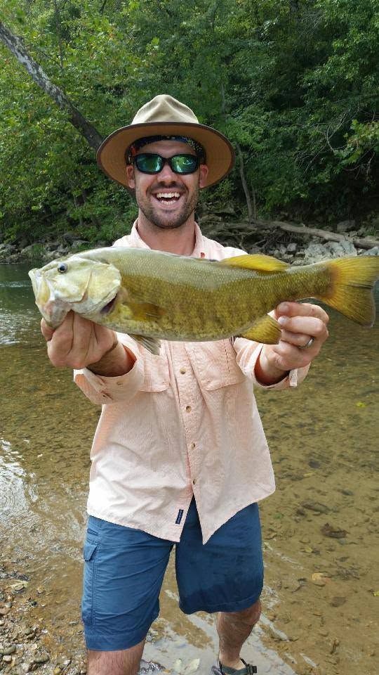 Man holding a large fish by a muddy river, wearing a straw hat, sunglasses, and a beige shirt.