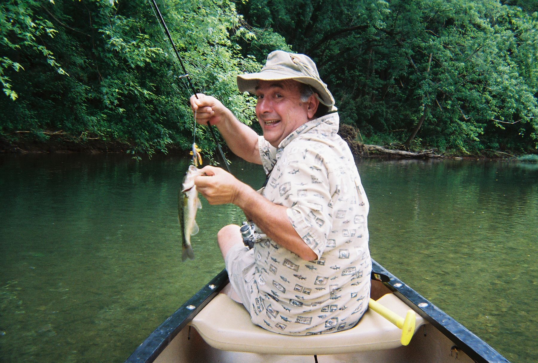 Person in a hat smiling while holding a fish in a canoe on a calm green river