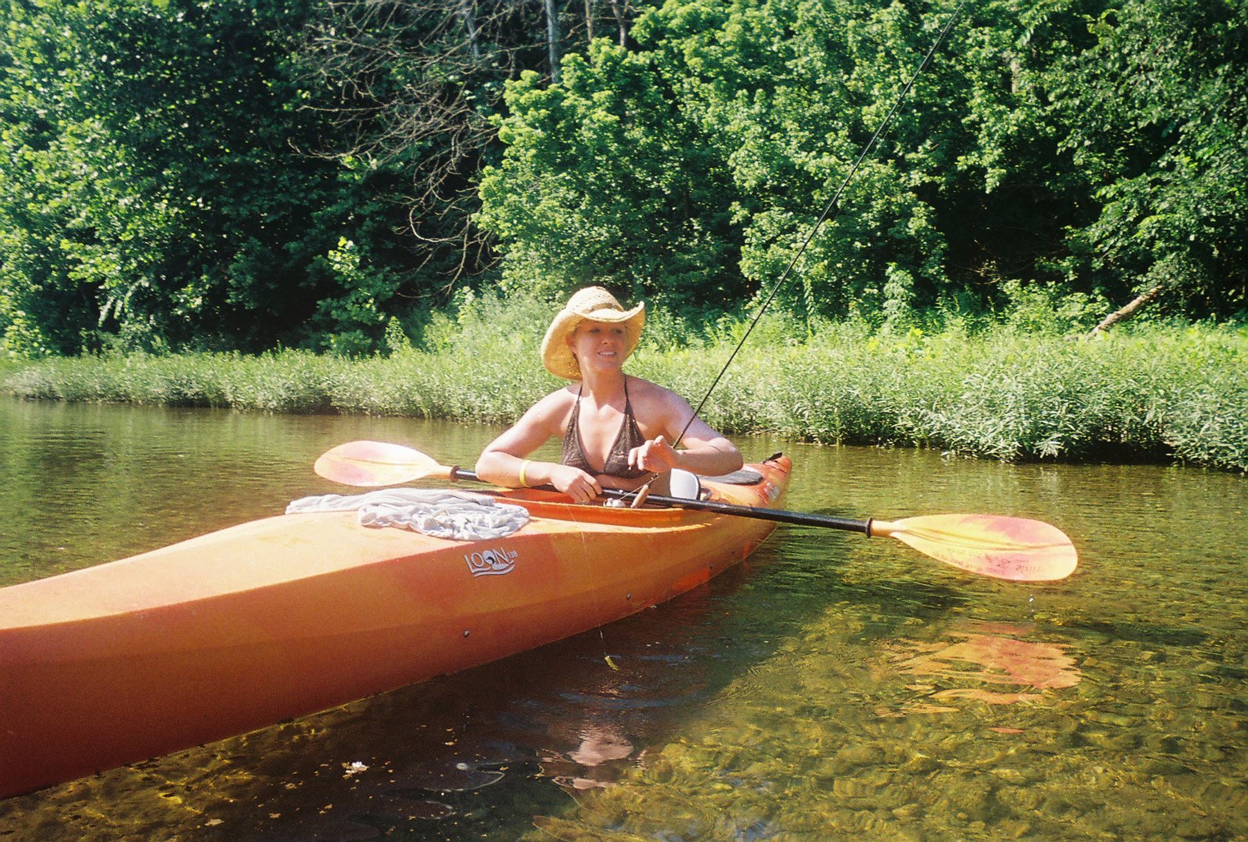 Person in a straw hat kayaking on a calm river with green trees along the bank