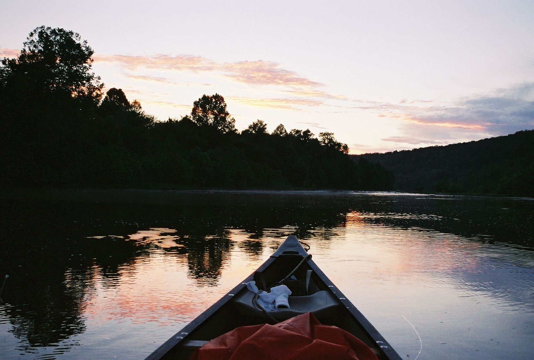 View from a canoe on a calm lake at sunset, with silhouetted trees and reflections on the water