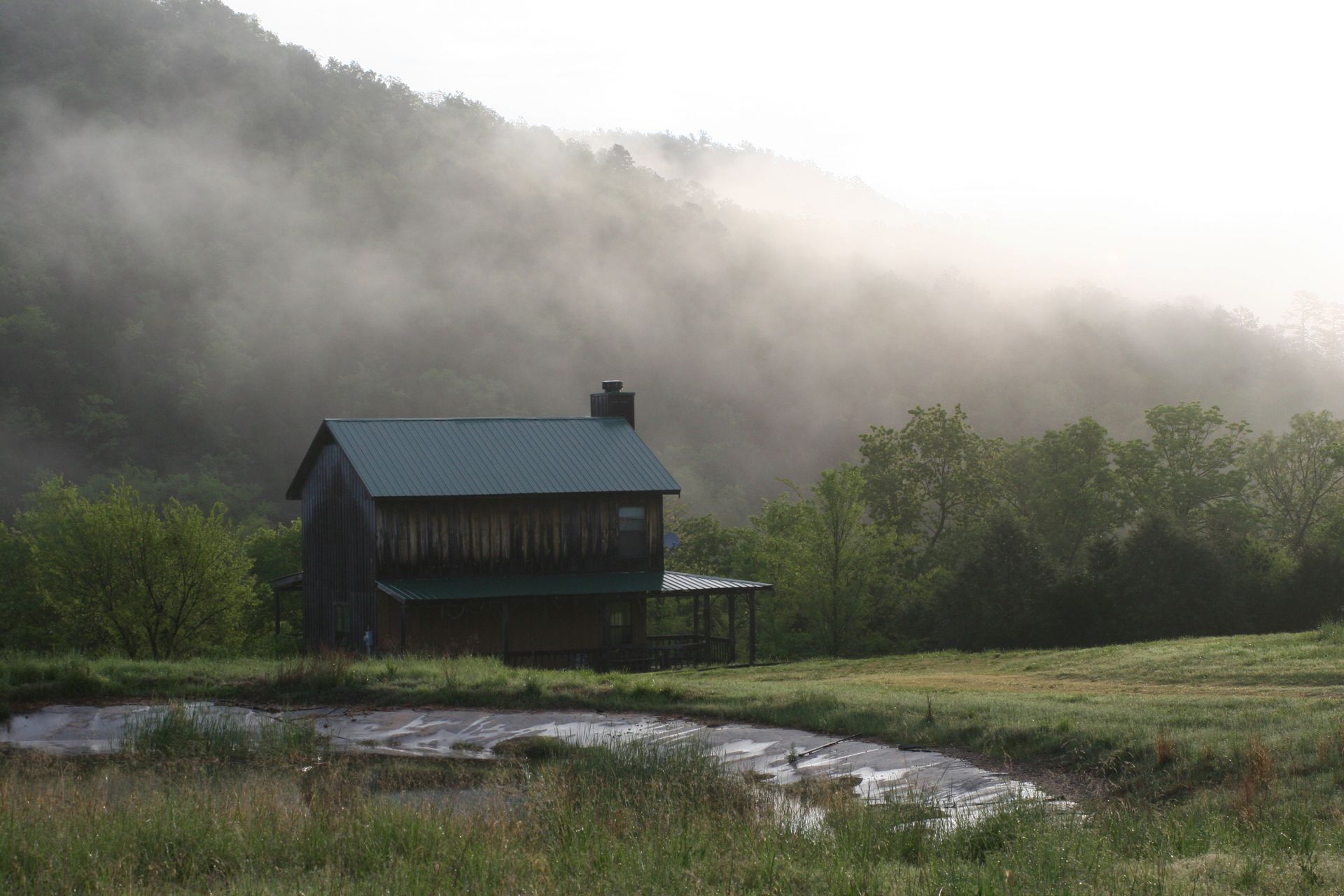 Small dark cabin in a misty green valley beside a winding stream at dawn