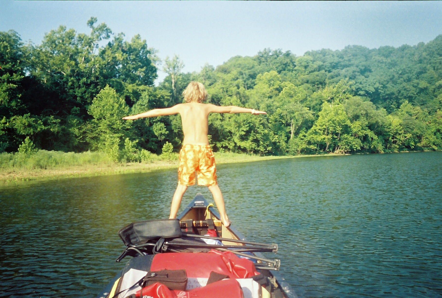 Person standing with arms outstretched on a red boat in a lake, with green trees and hills behind