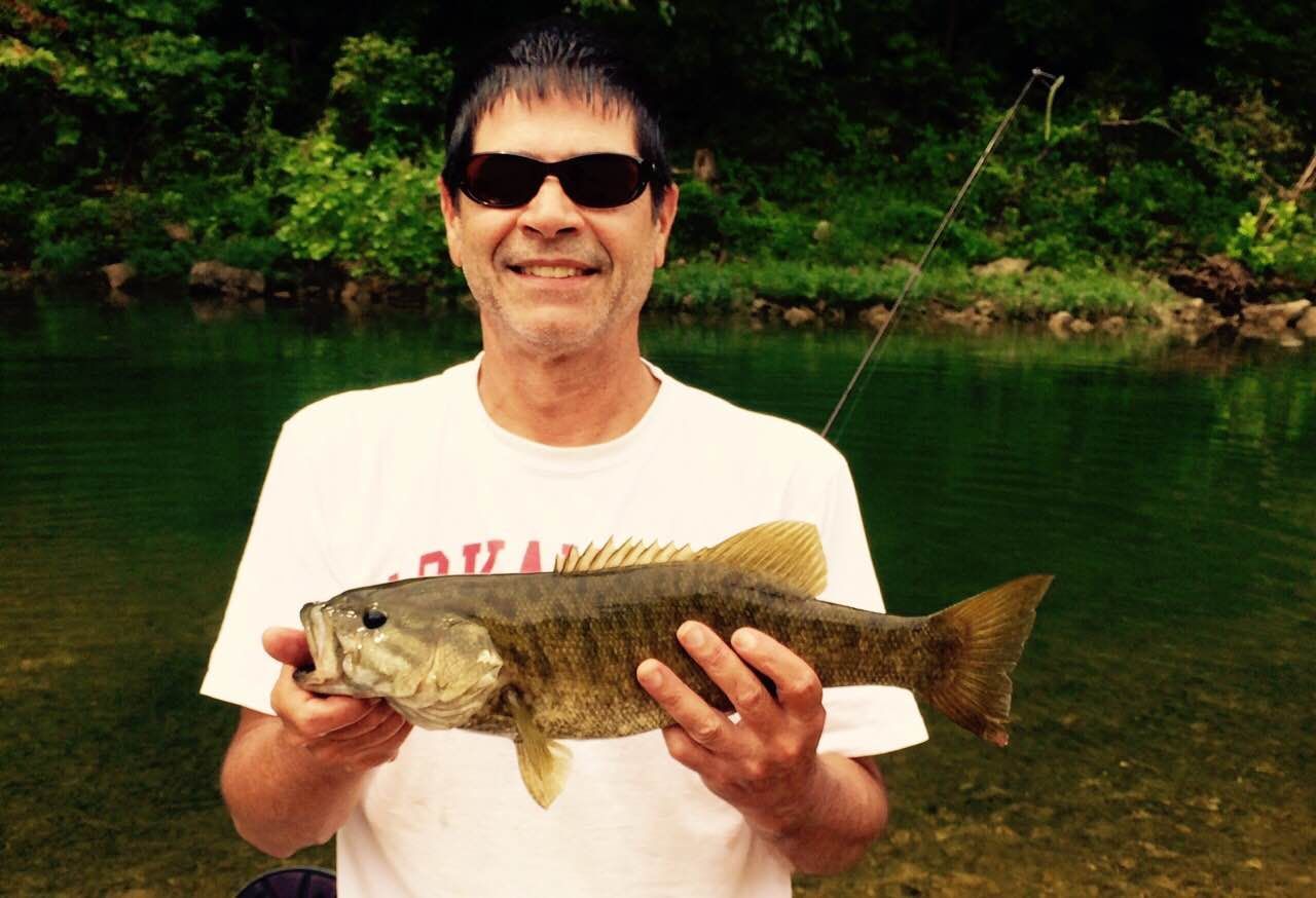 Smiling man in sunglasses holding a large fish by a green river