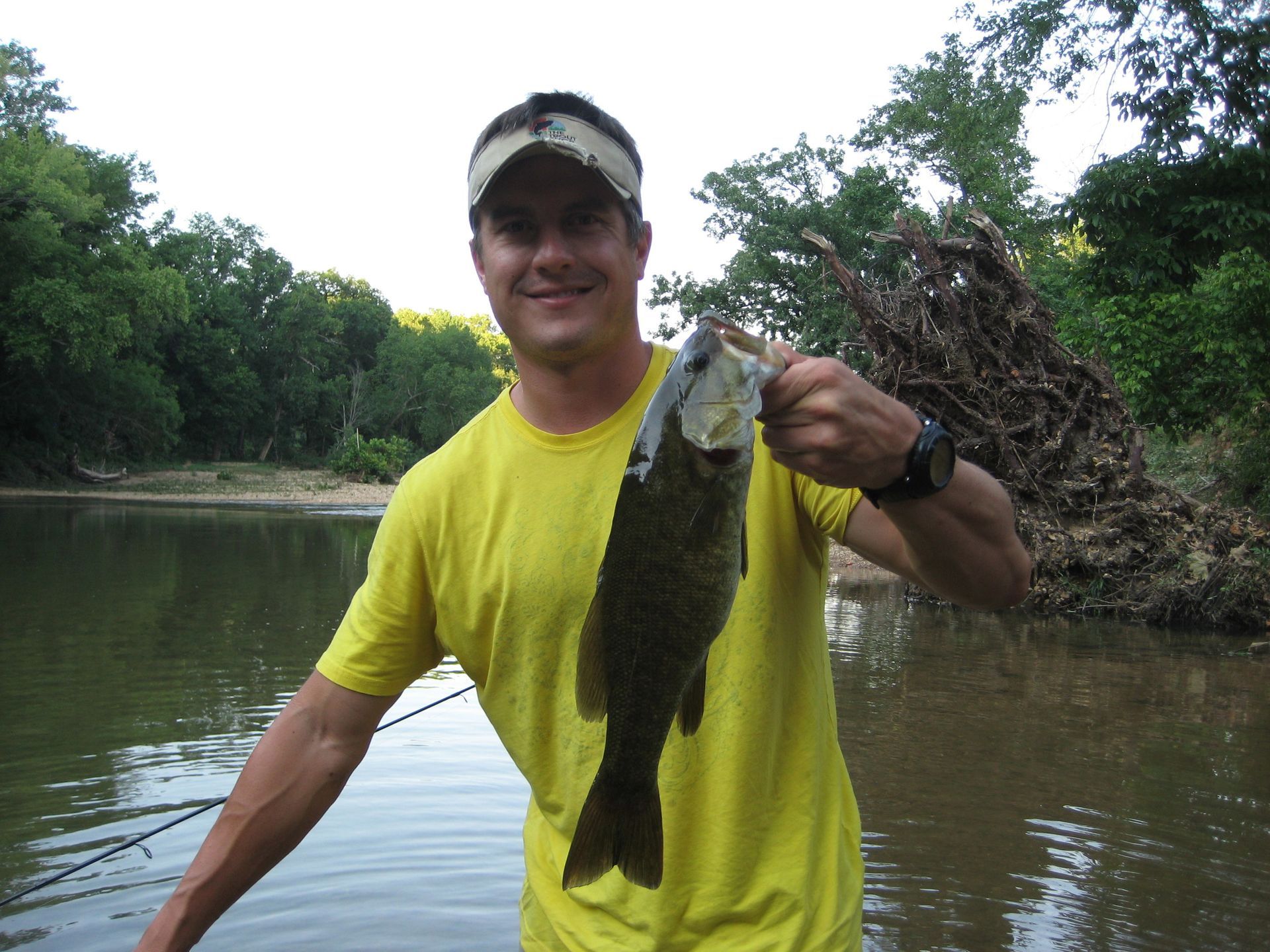Smiling fisherman in yellow shirt holding a large fish by a river