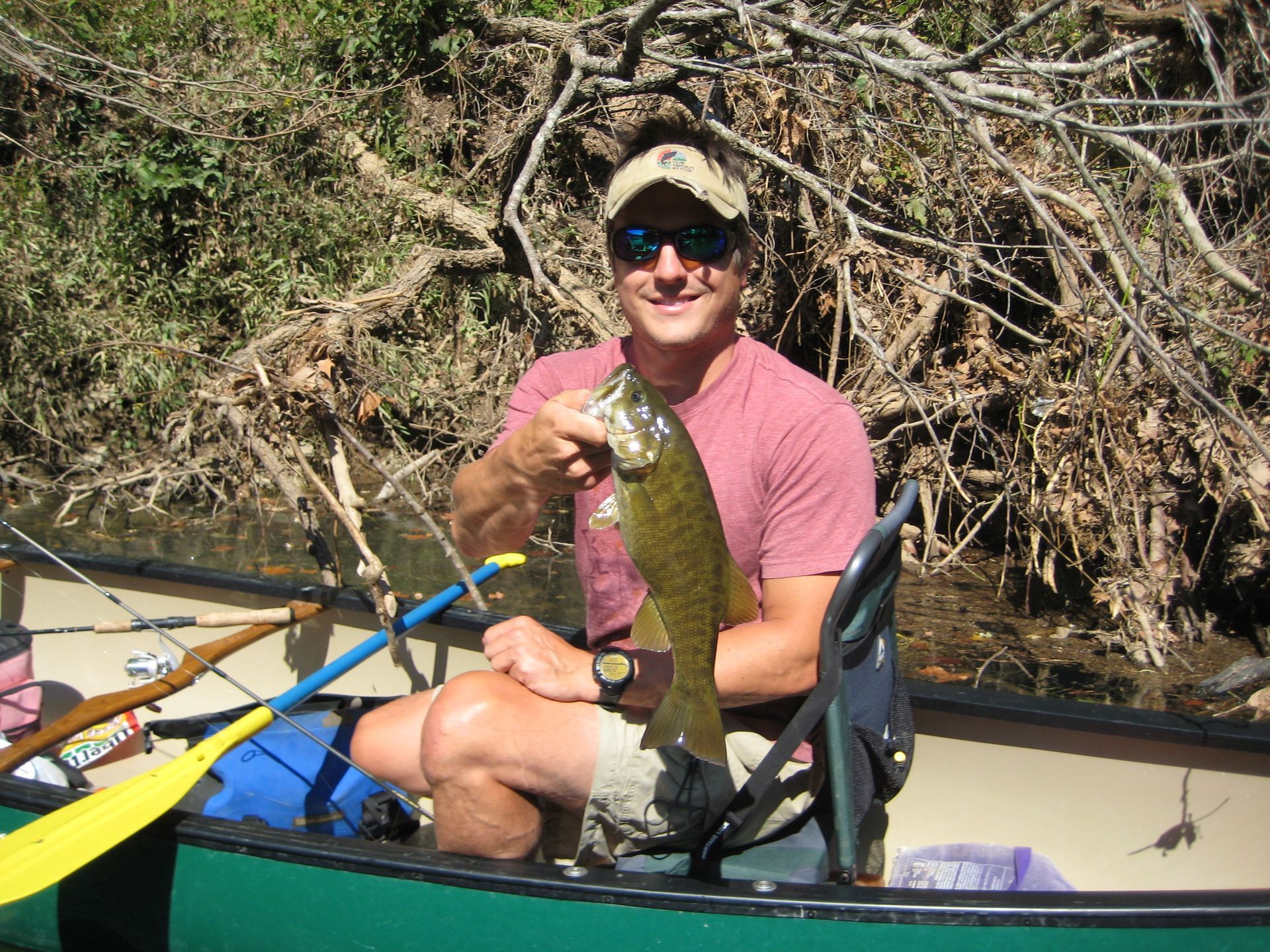 Kayaker in a canoe holds up a fish beside a riverbank with tangled brush.