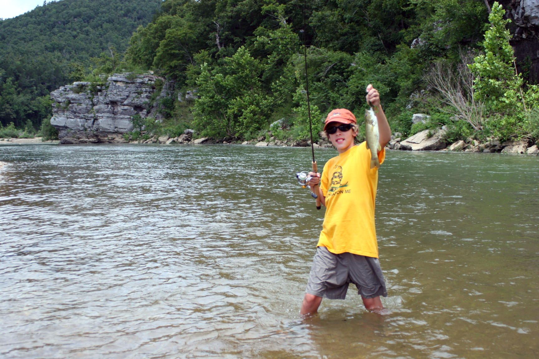 Person standing in a river, holding up a fish and fishing rod, with wooded cliffs in the background