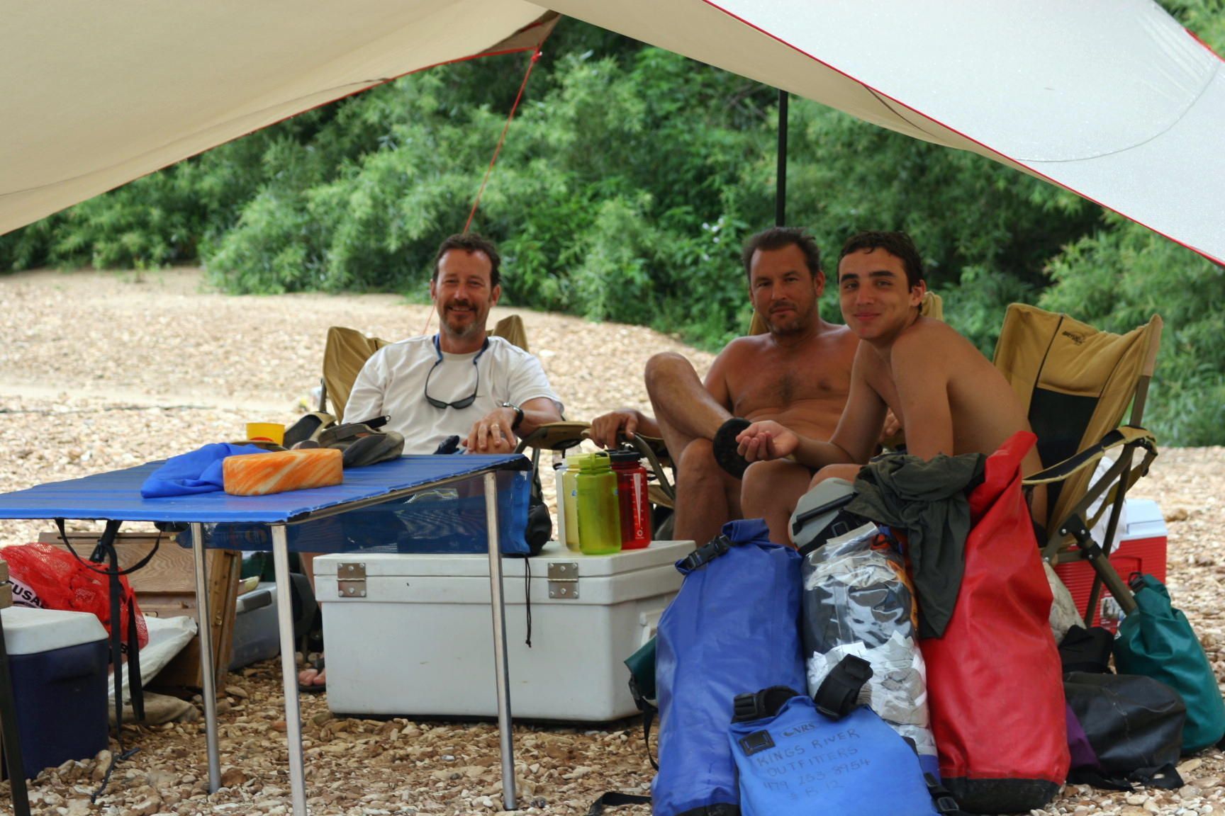 Three people relaxing under a canopy at a campsite beside coolers and camping gear.