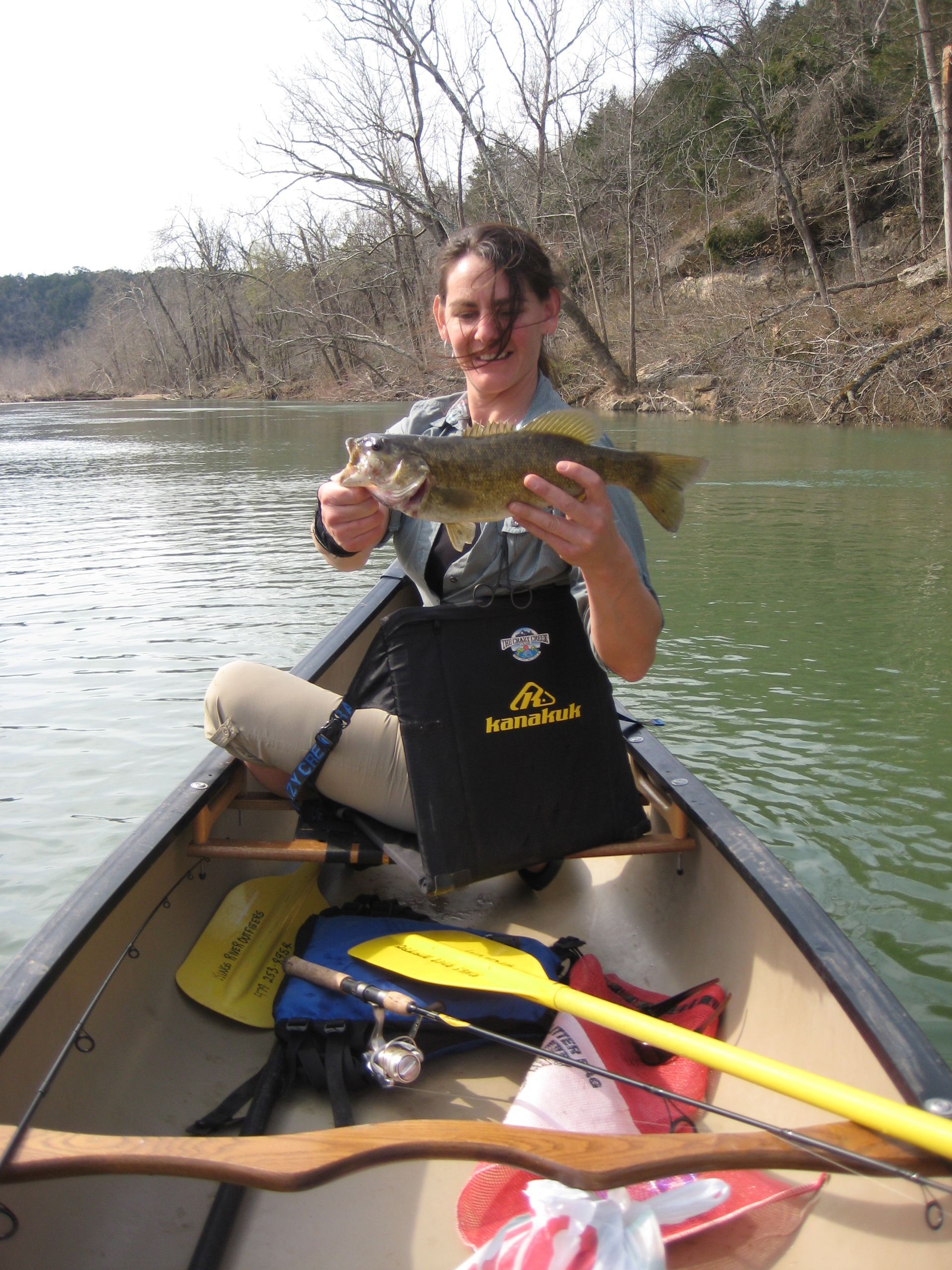 Person smiling in a canoe, holding a fish on a calm river, with trees along the shore.