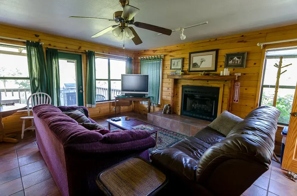 Cozy living room with leather sofas, fireplace, TV, ceiling fan, and large windows overlooking greenery