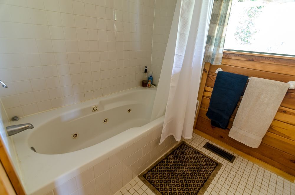 Bathroom with white tub, tiled floor, and towels hanging by a windowed wooden wall.
