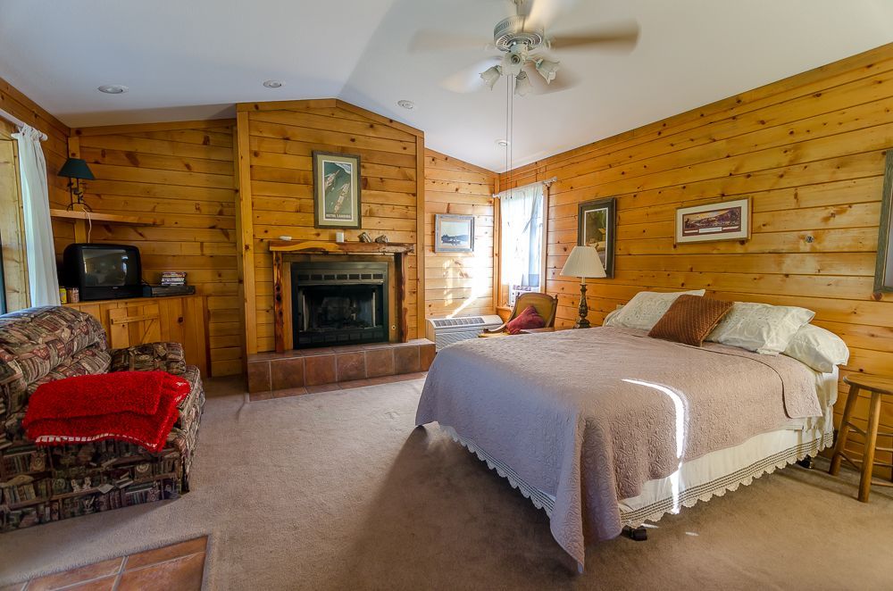 Cozy wood-paneled bedroom with fireplace, queen bed, red chair, and ceiling fan