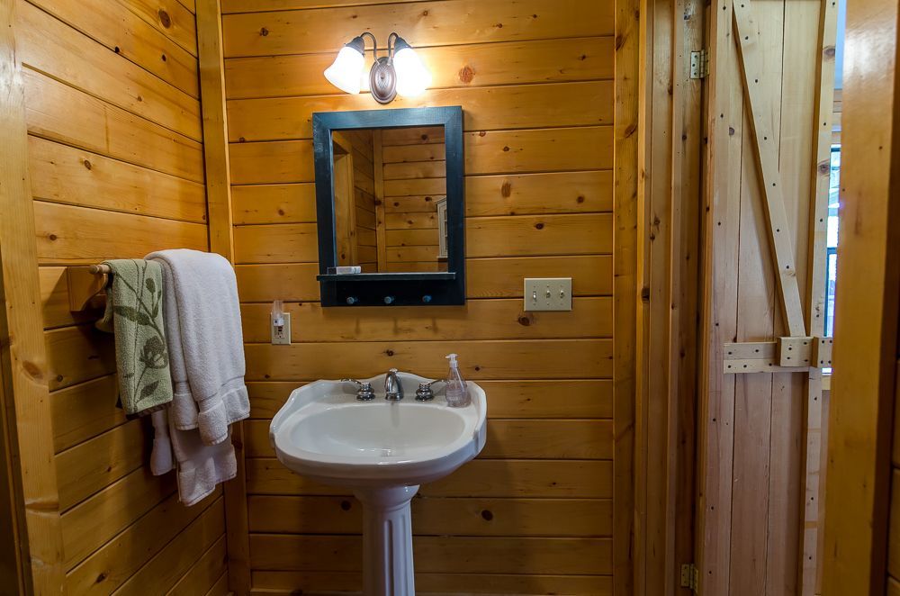 Rustic wood-paneled bathroom with pedestal sink, mirror, light, and towels hanging on the wall