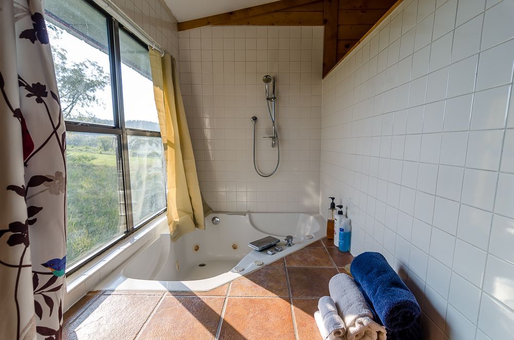 Bathroom with white tiled walls, corner bathtub, shower, window, and towels on a terracotta floor.