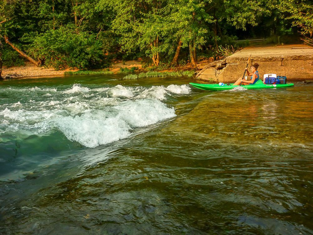 Two people in a green raft riding whitewater near a sandy riverbank and forested shore