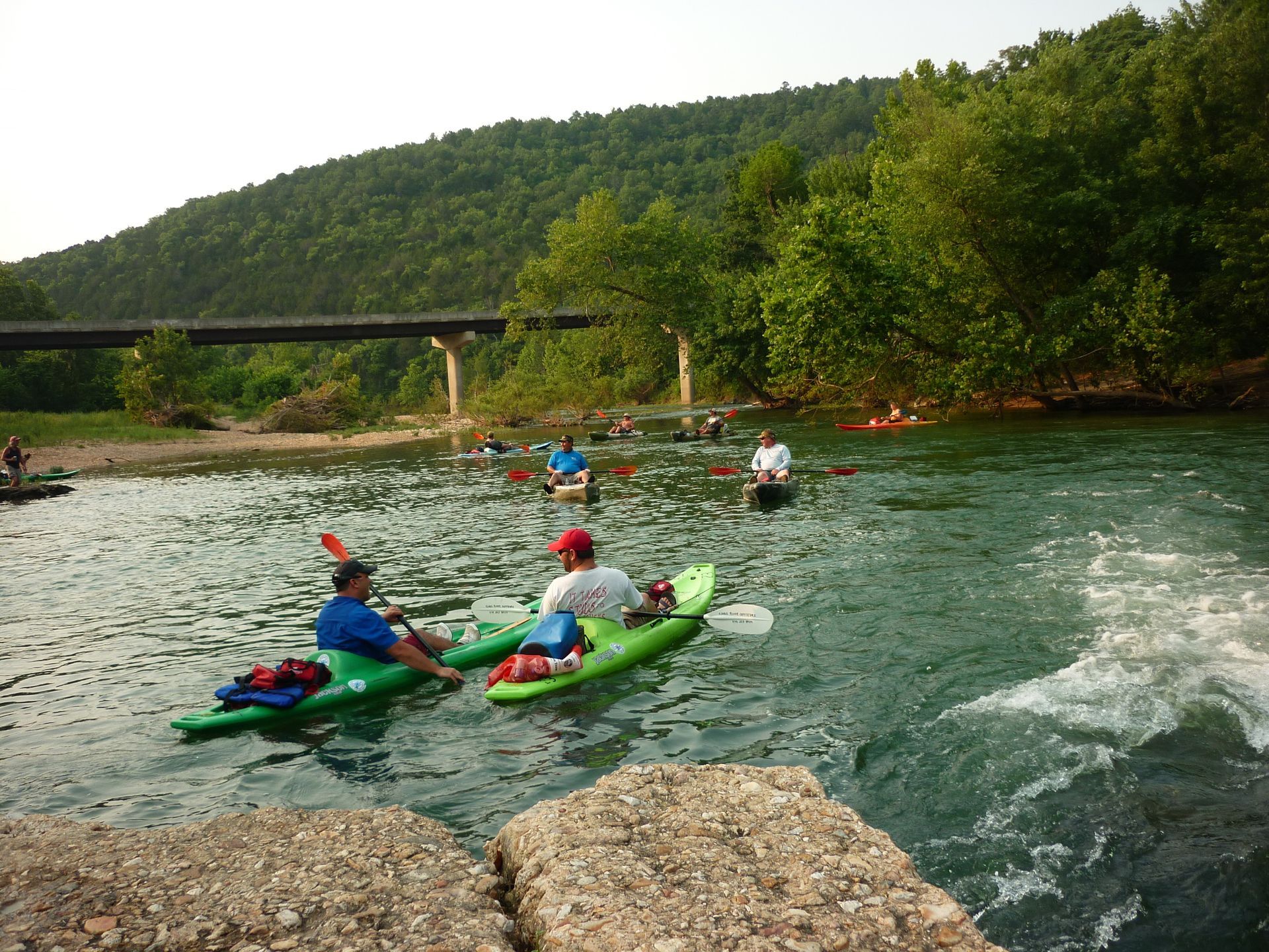 Kayakers in colorful boats paddling on a river near rocks, trees, and a bridge