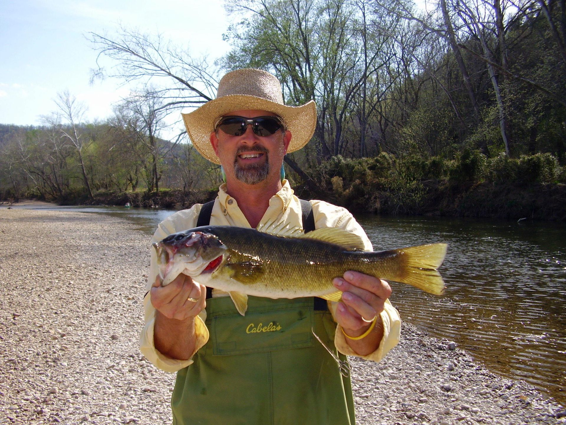 Angler in a straw hat holding a large fish by a riverbank