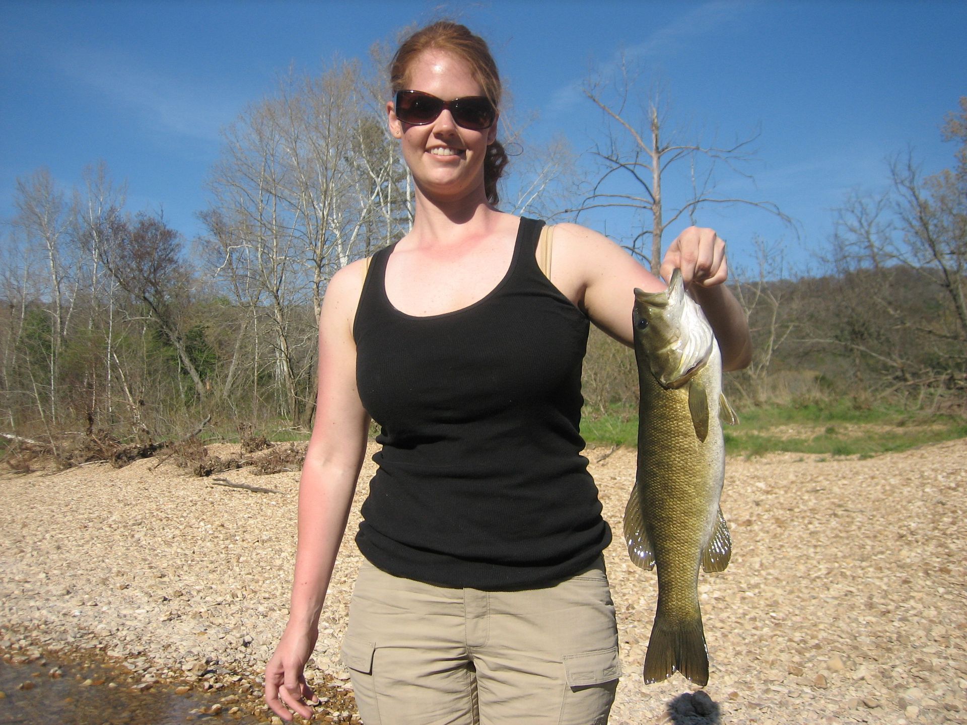 Person holding a fish on a sunny rocky beach, wearing sunglasses and a black tank top.