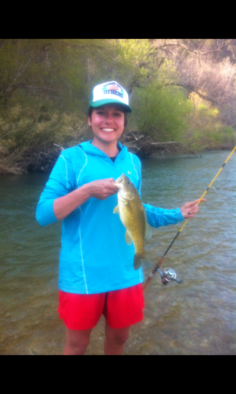 Person smiling while holding a fish by a river, wearing a blue shirt, red shorts, and a cap.