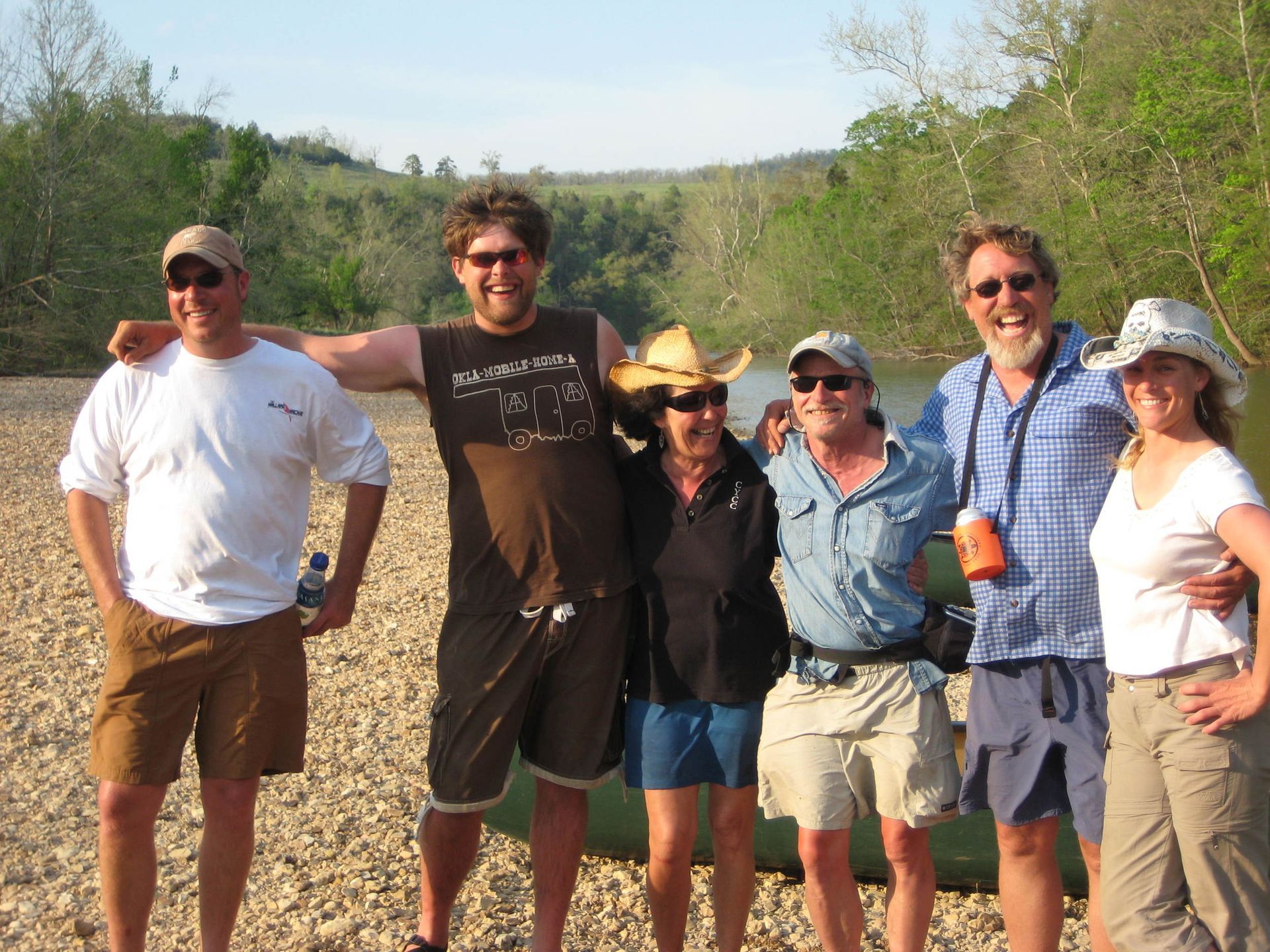 Six hikers smiling outdoors on a trail, with trees and hills in the background.
