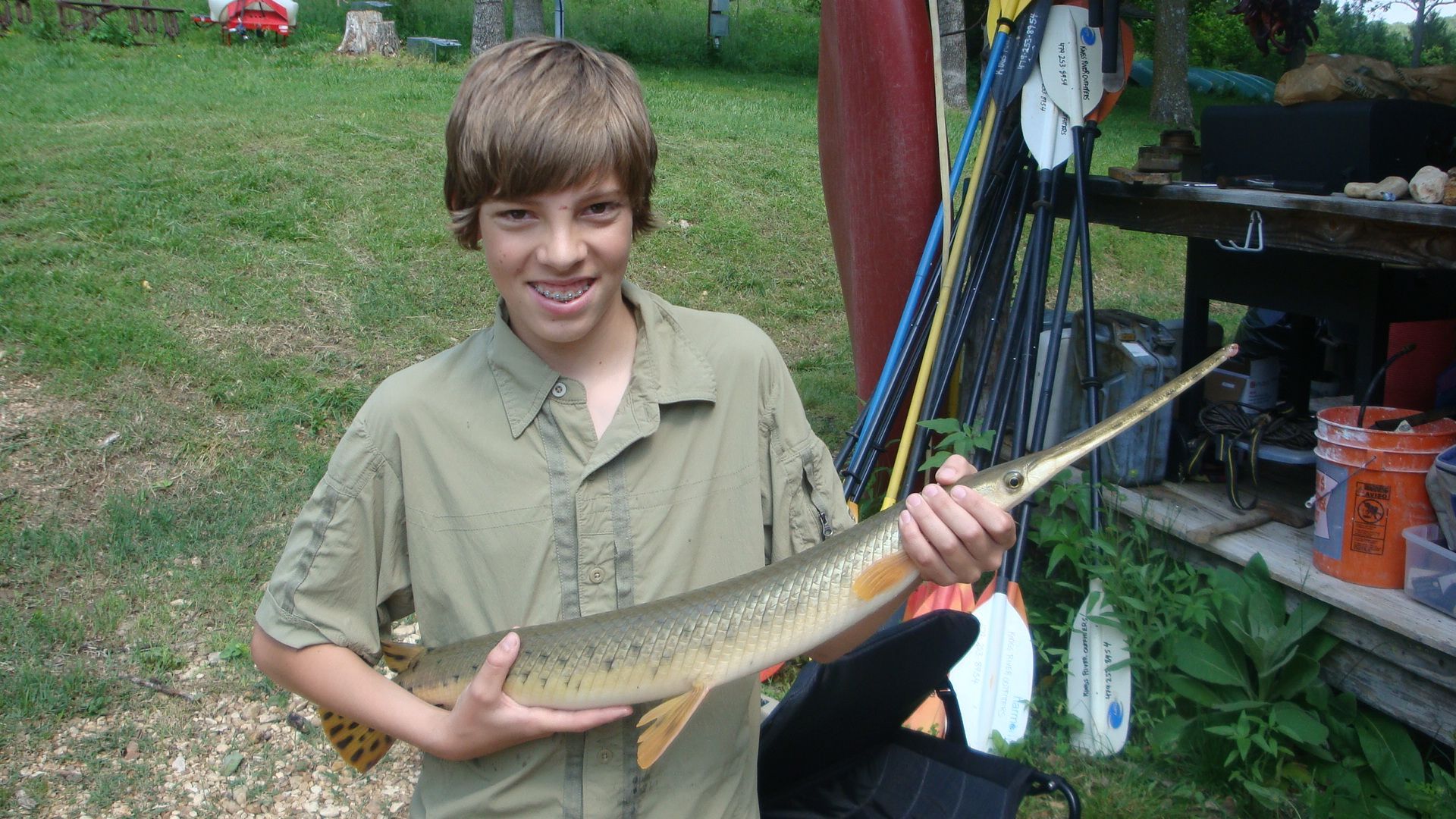 Smiling boy holding a long fish outdoors beside a shed and fishing gear.