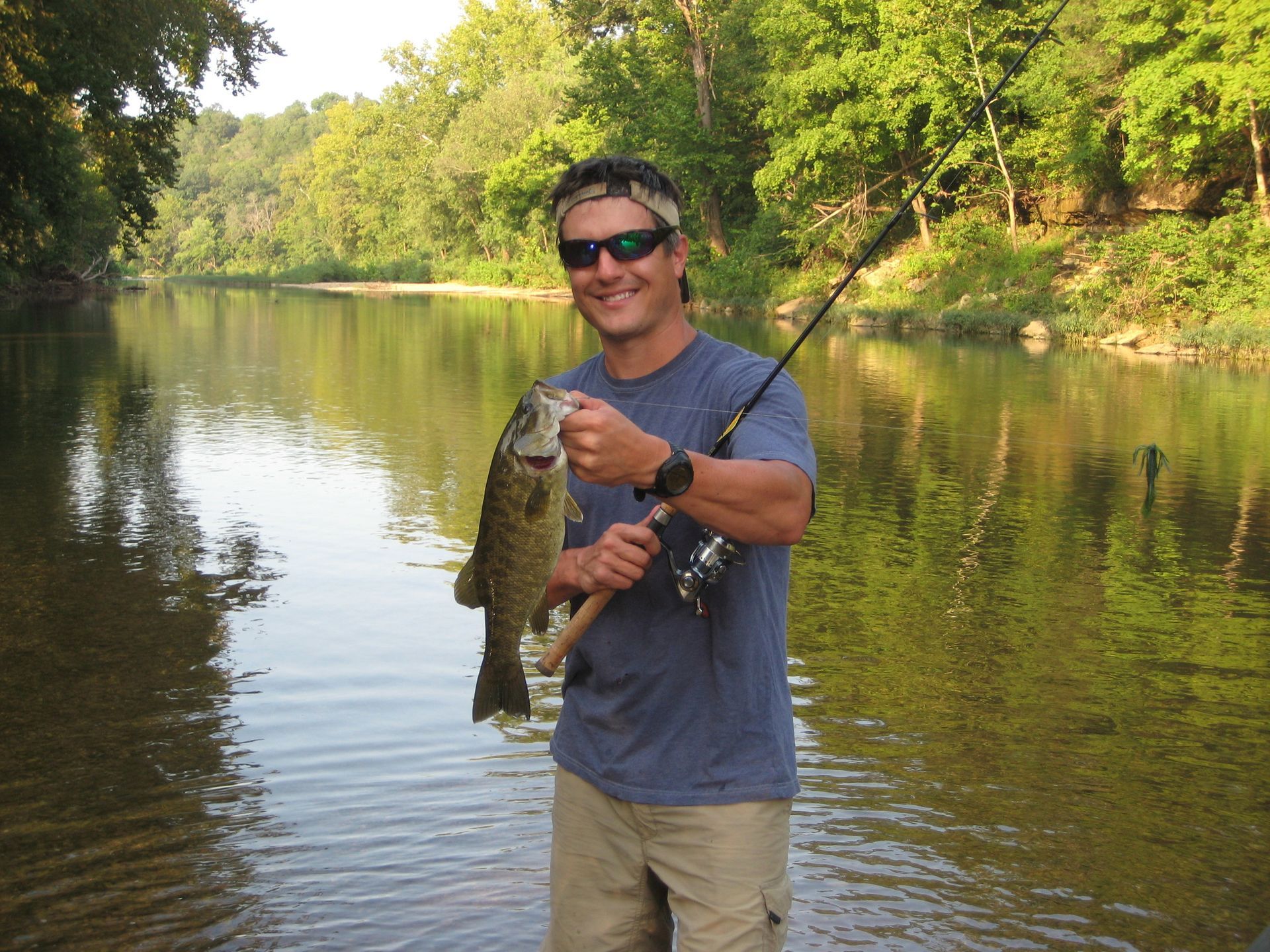 Man holding a large fish by a calm river, wearing sunglasses and a blue shirt.