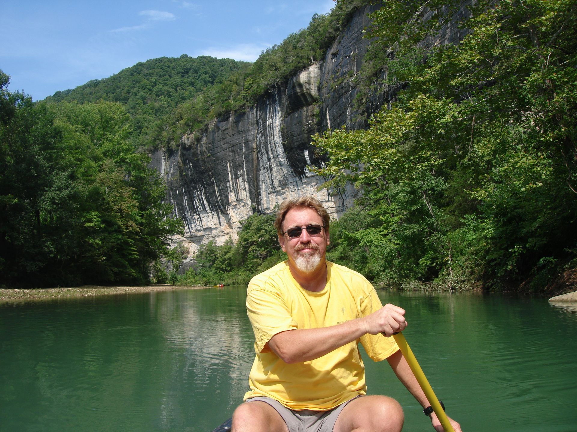 Man in a yellow shirt paddling a canoe on a green lake beside rocky cliffs and trees