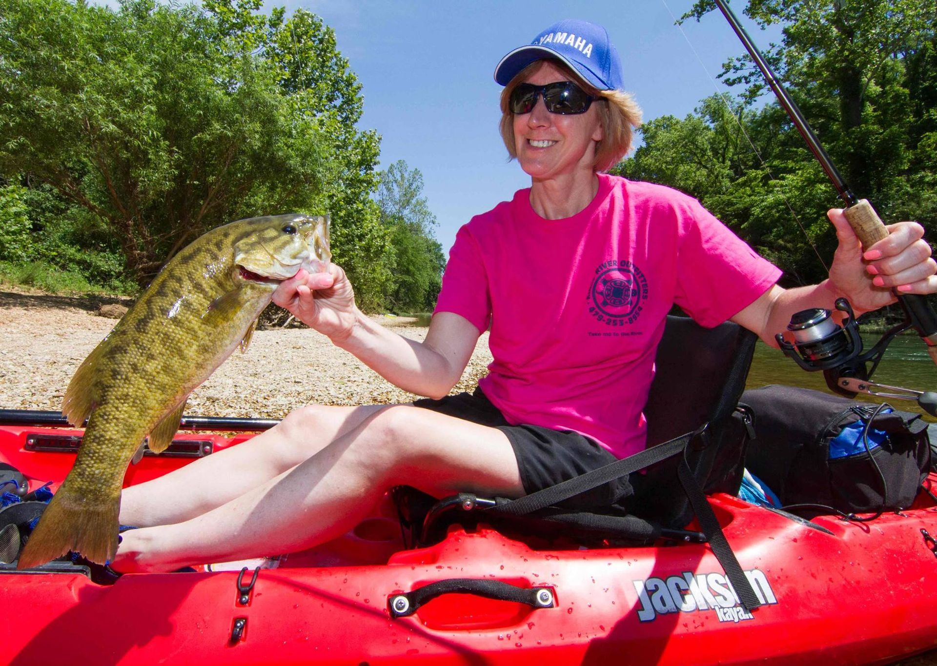 Angler in pink shirt holding a fish while sitting in a red kayak on calm water