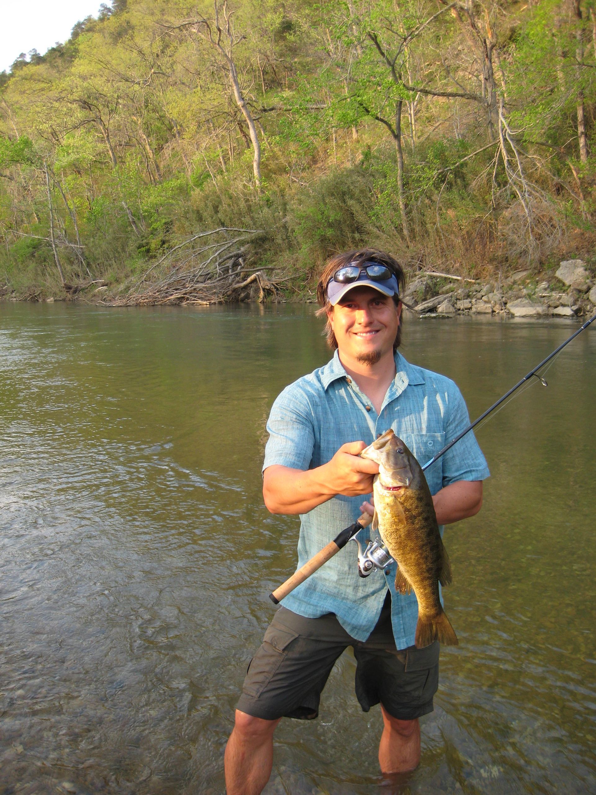 Person standing in a river, smiling and holding a fish while fishing near a wooded bank.