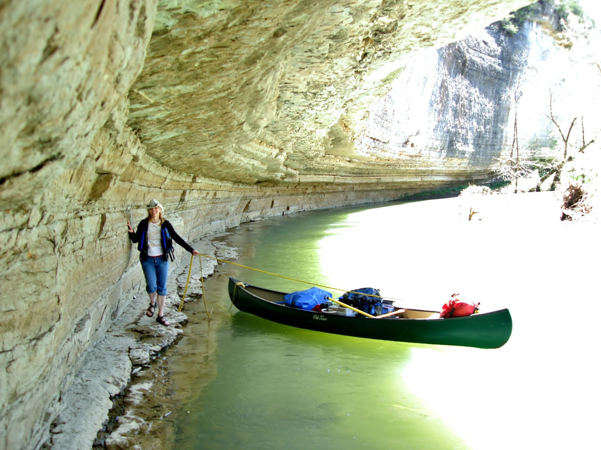 Person standing beside a green canoe in a rocky river cave with bright light in the distance