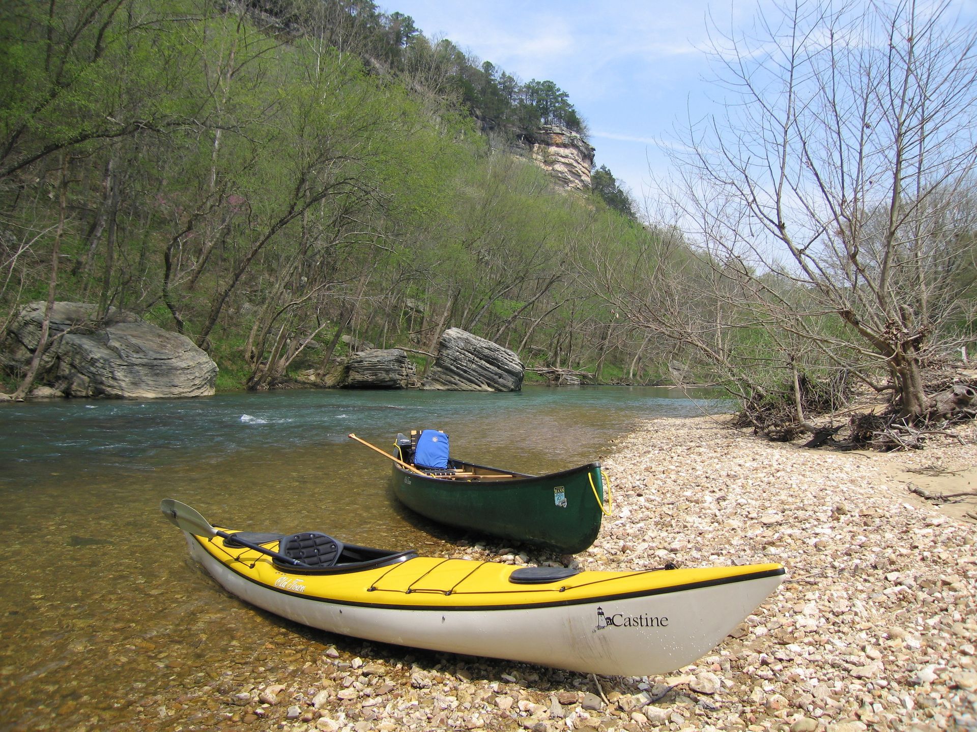 Two canoes on a rocky riverbank, one green and one yellow, beside a calm river and wooded hillside.