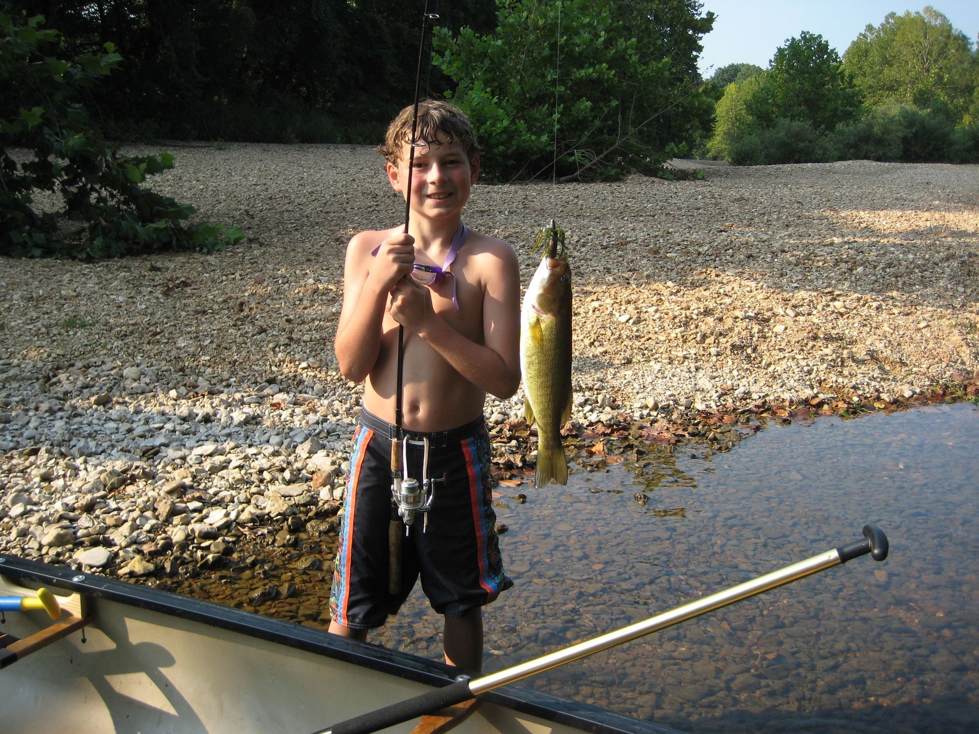 Child holding a fish beside a canoe in a shallow riverbed, smiling outdoors.
