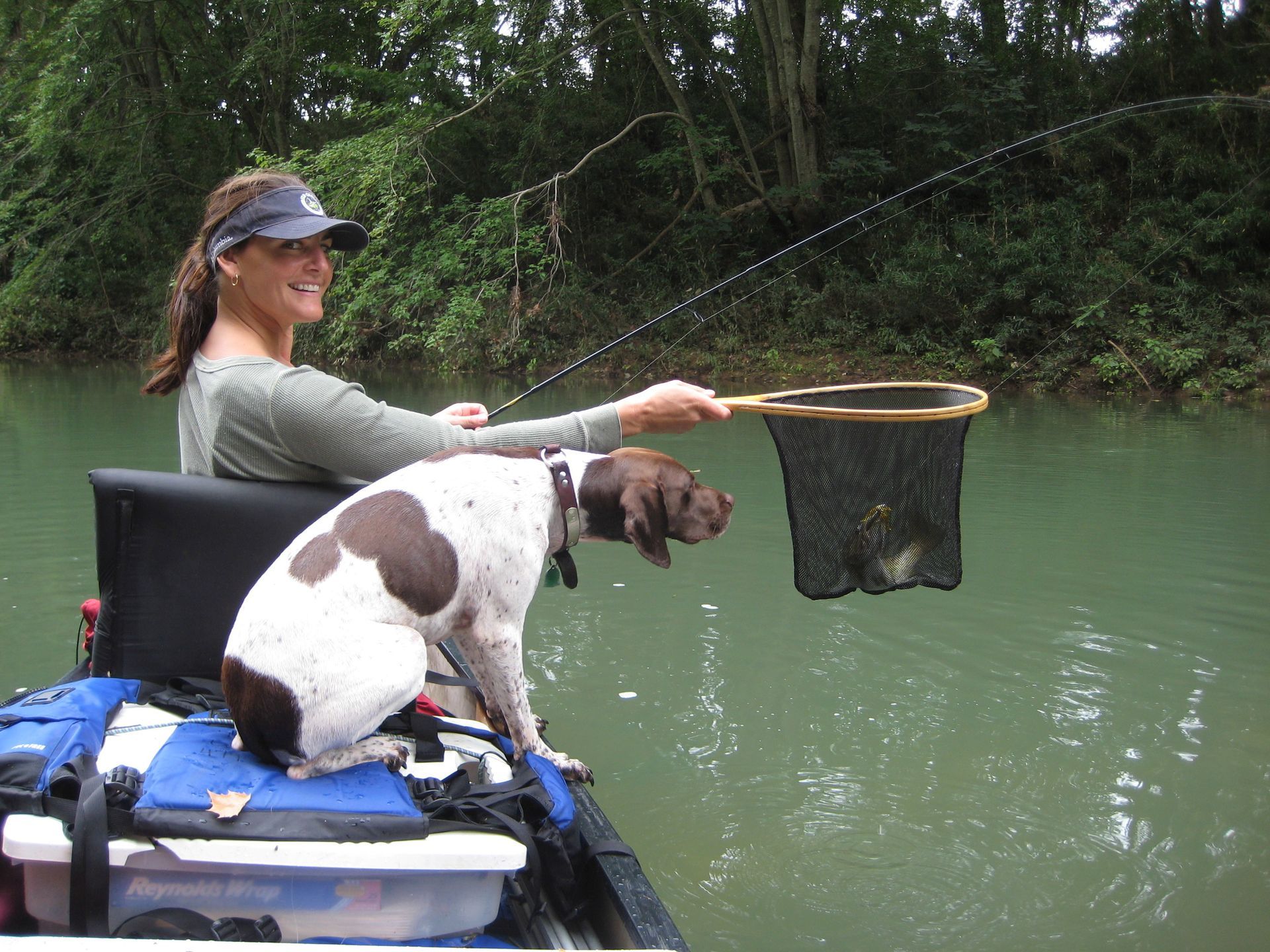 Person fishing from a kayak with a dog on board, using a net over calm green water.