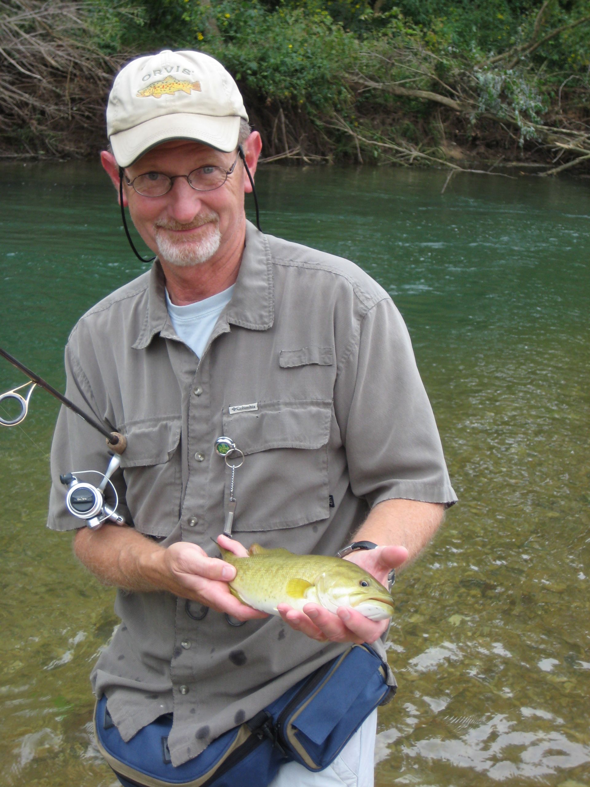 Man holding a small fish beside a river, wearing a cap and fishing vest.