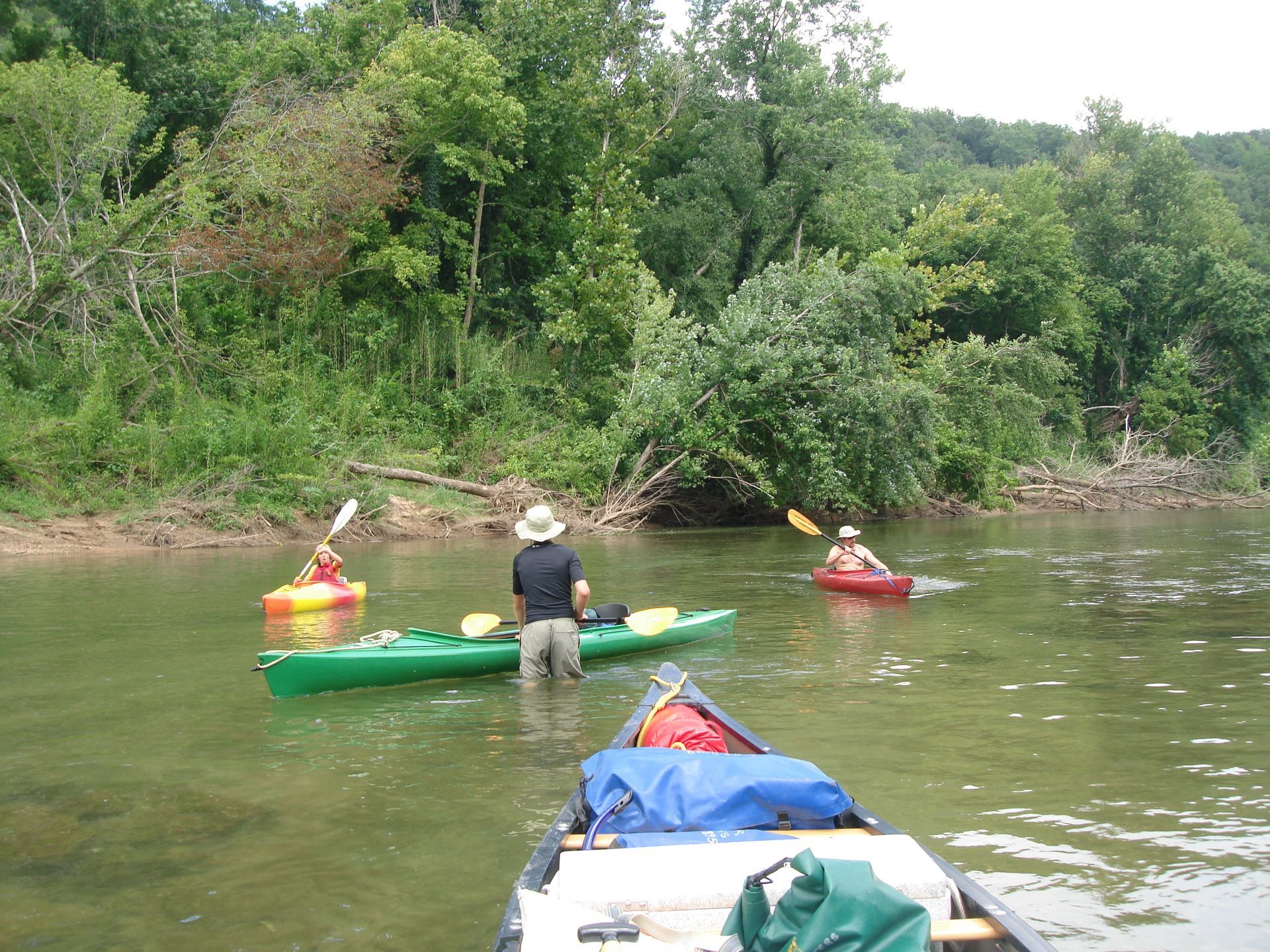 People in colorful kayaks on a calm river beside a wooded bank