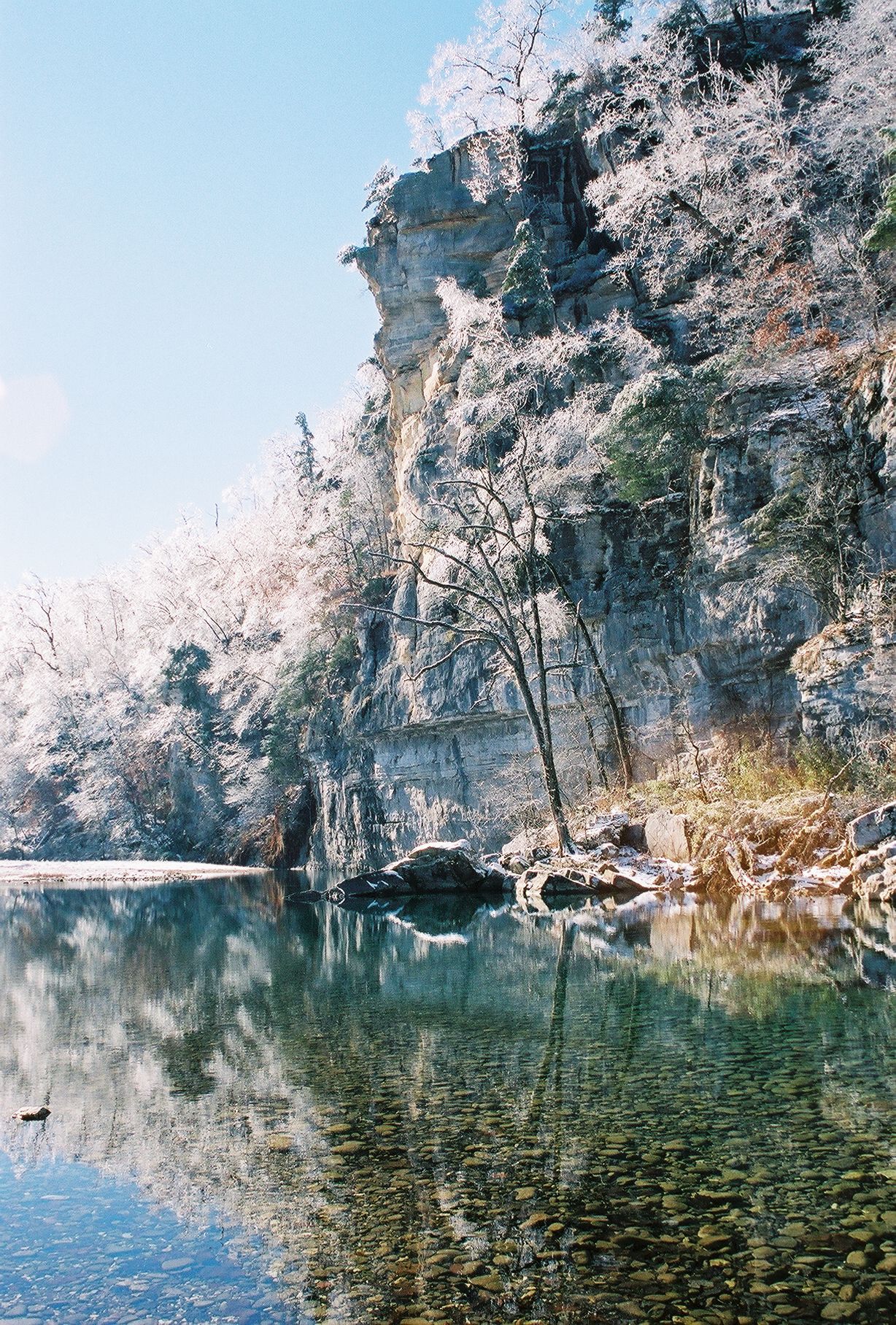 Snow-covered cliffs and trees reflected in a calm lake under a blue sky