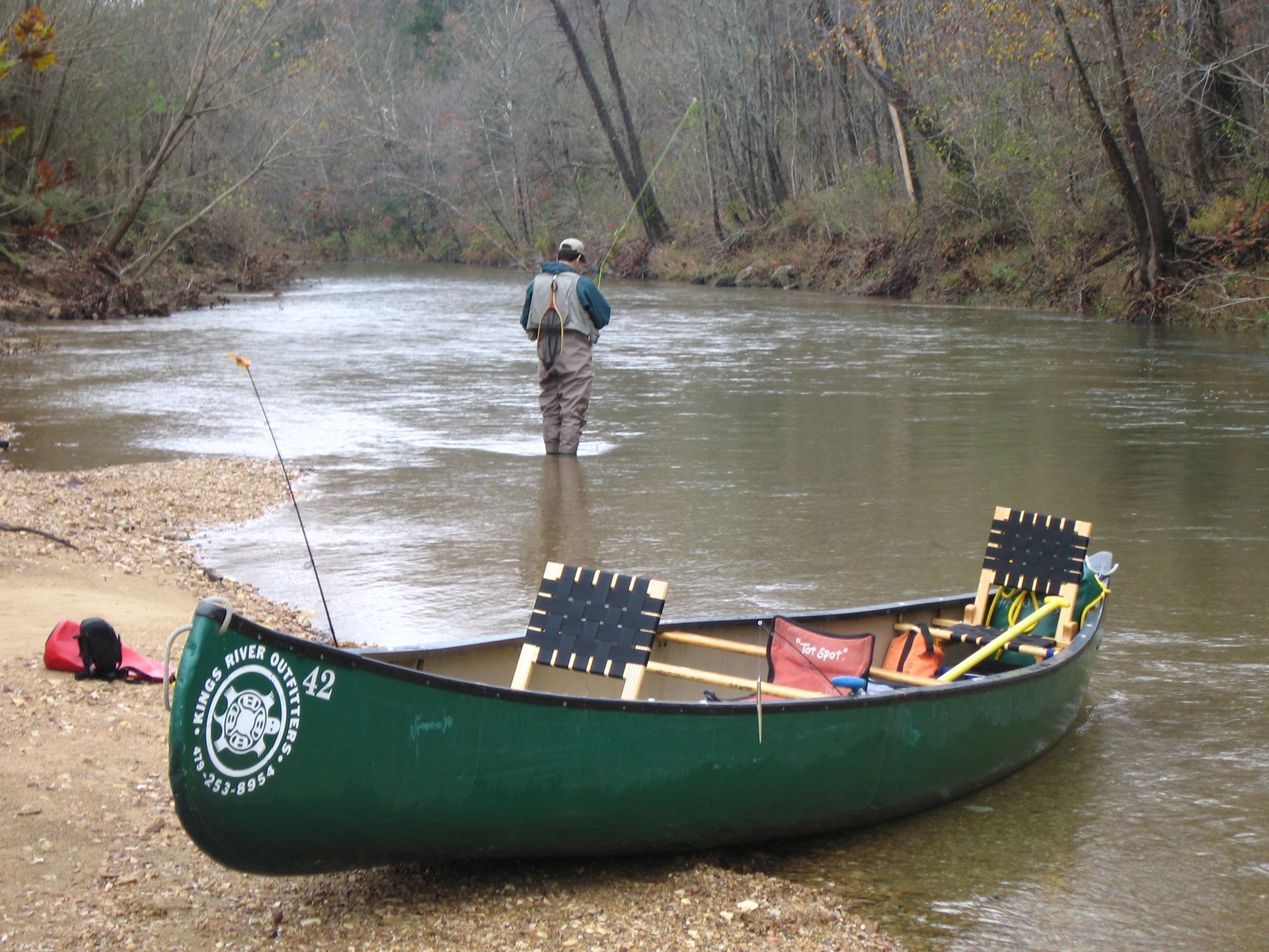 Green canoe on a sandy bank beside a river, with a person wading in the water nearby.