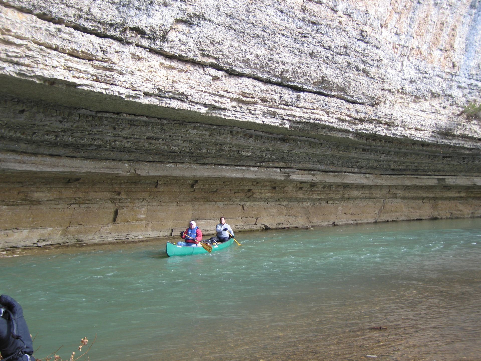 Two people in a turquoise kayak on calm water beneath a layered rock cliff