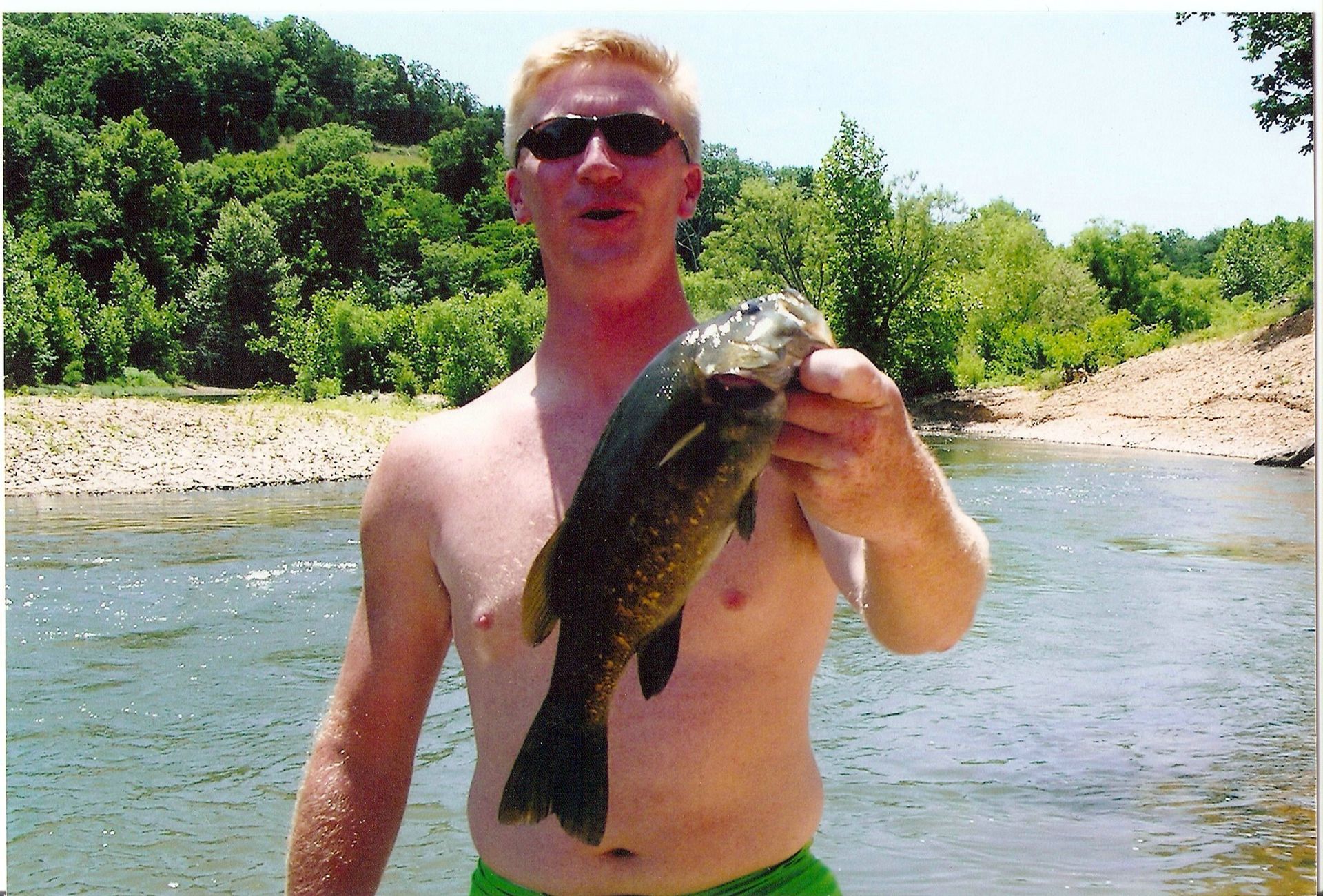 Man holding a fish in a river, wearing sunglasses and green shorts, with trees and rocks in the background