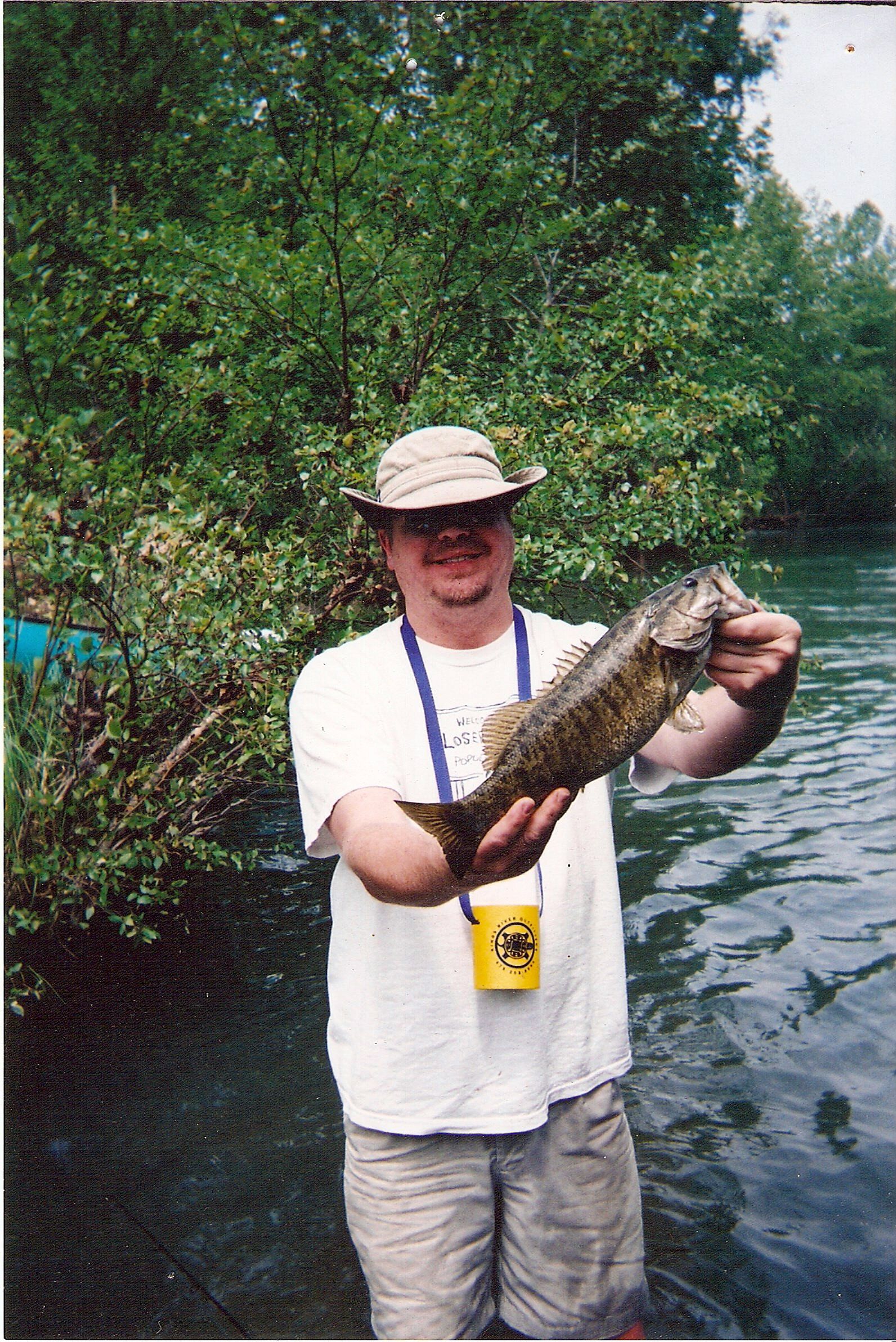 Man holding a fish in a river, wearing a hat and white shirt, with green trees in the background