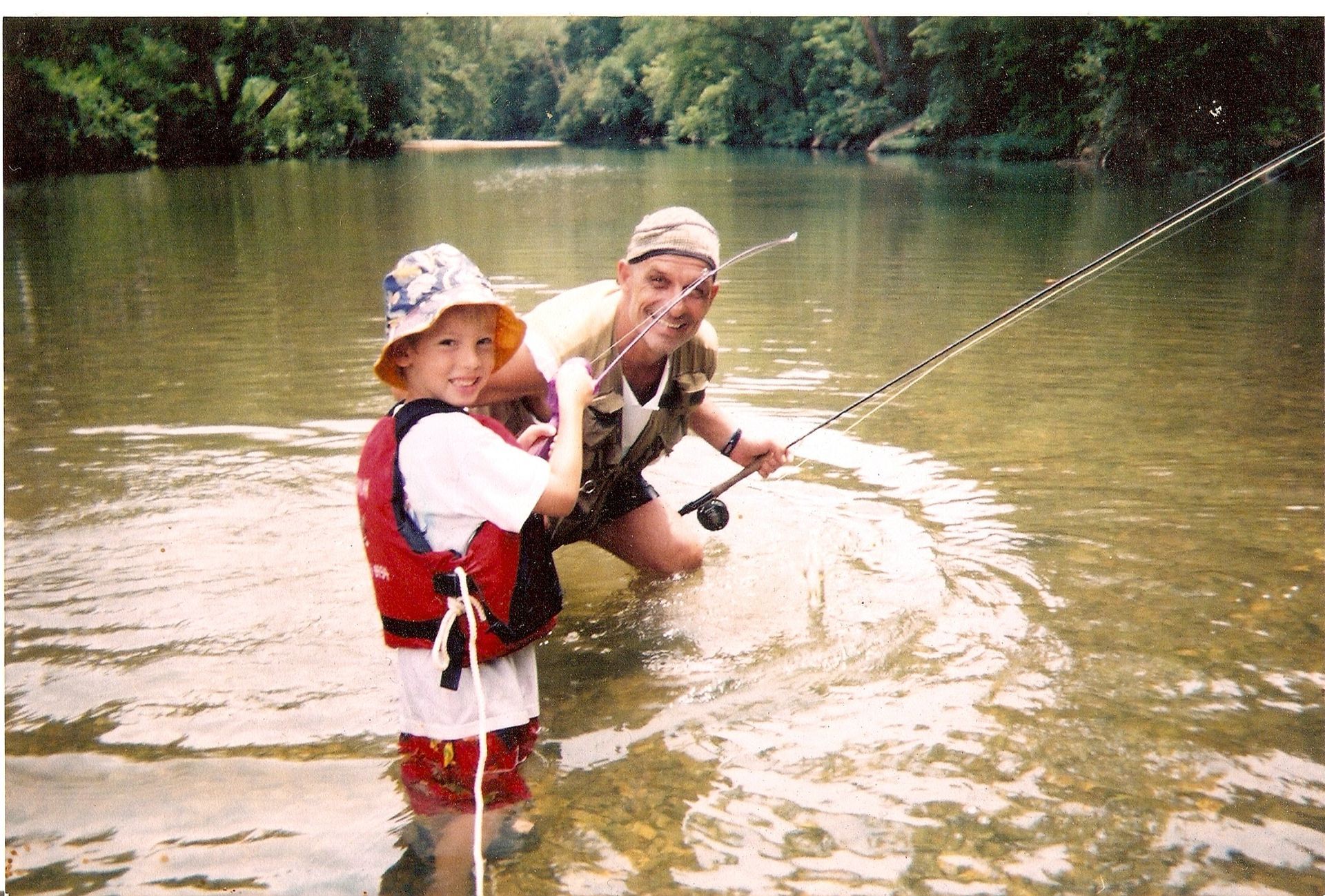 Two people fishing in a shallow river, one holding a rod and the other standing nearby in a life vest.