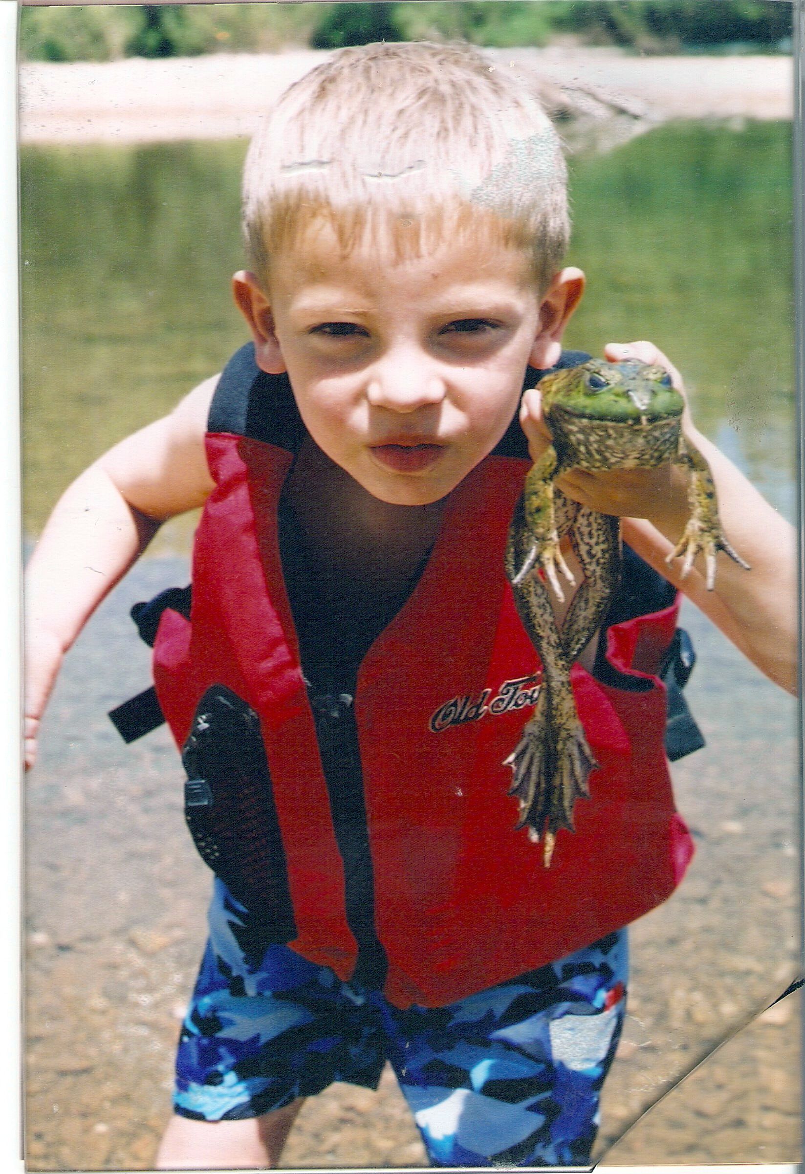 Young child in a red life vest holding up a fish outdoors near water