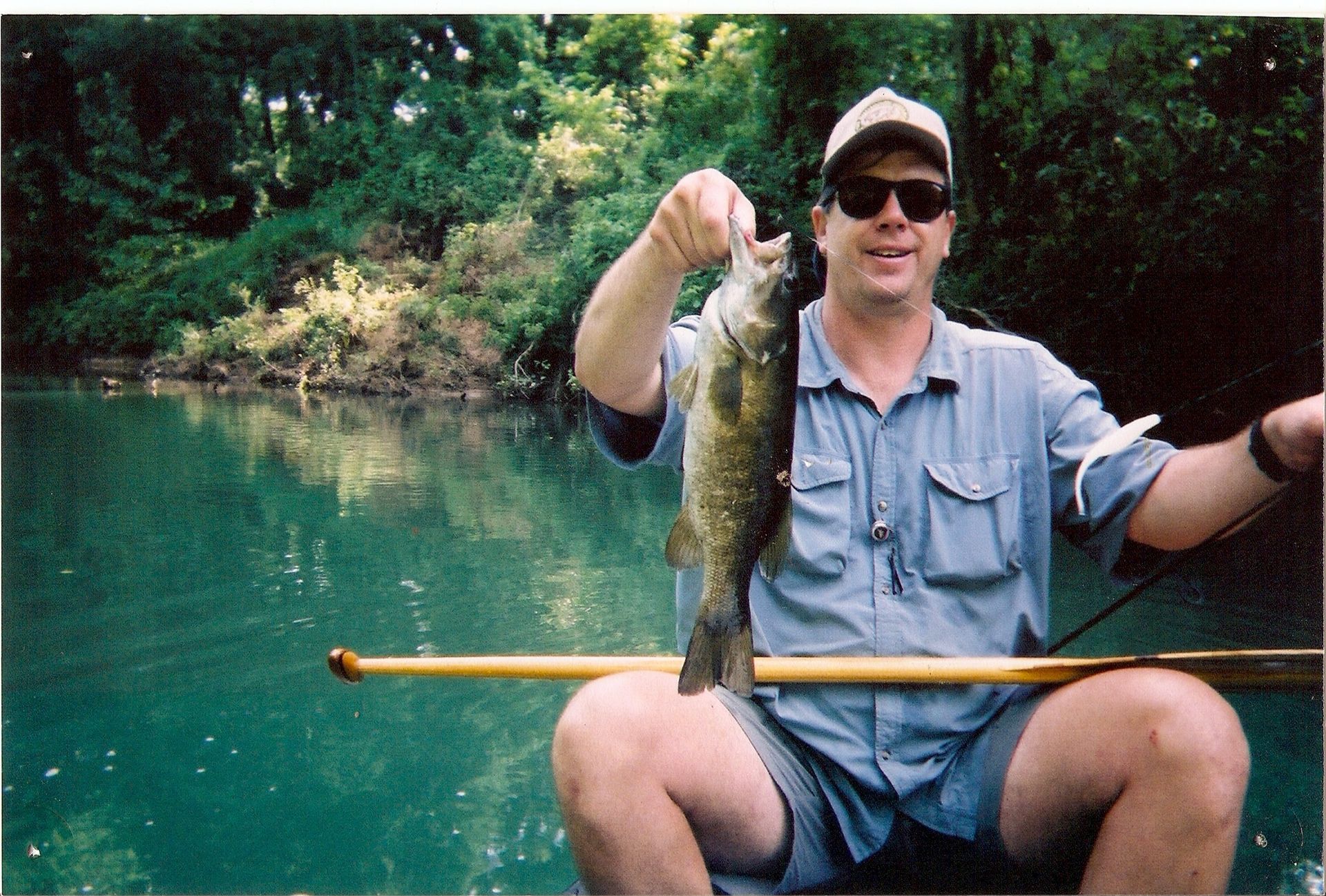 Man holding a fish over a net while seated in a canoe on a green lake, surrounded by trees