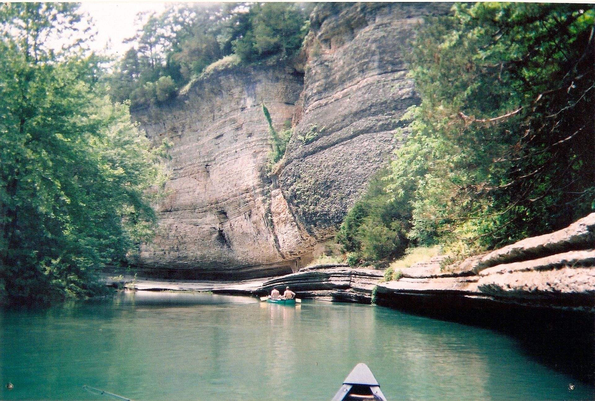 River beside rocky cliff with green trees and a canoe bow at the bottom center