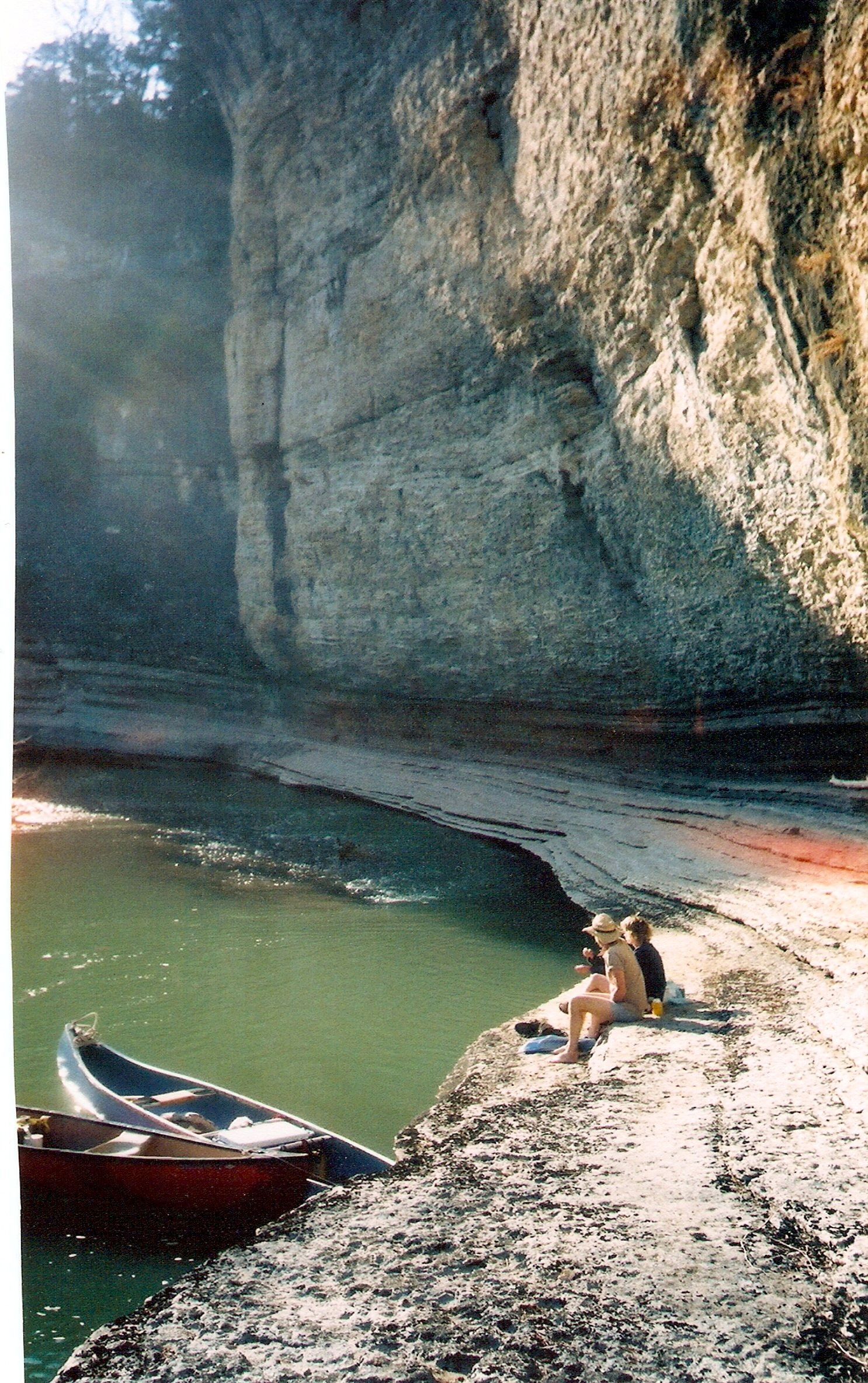 Cliffside beach cove with green water, pebbled shore, two people, and a red boat near the edge