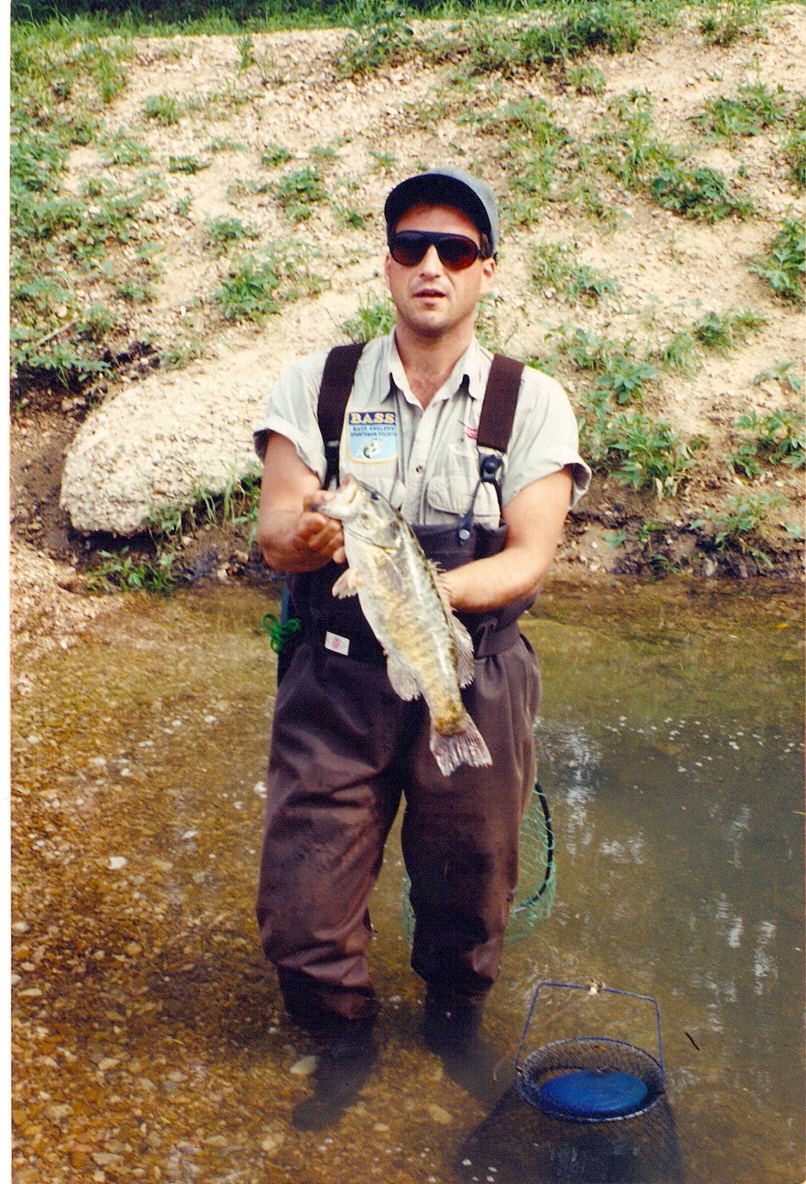 Person holding a fish while standing in a shallow stream, wearing a cap and sunglasses.